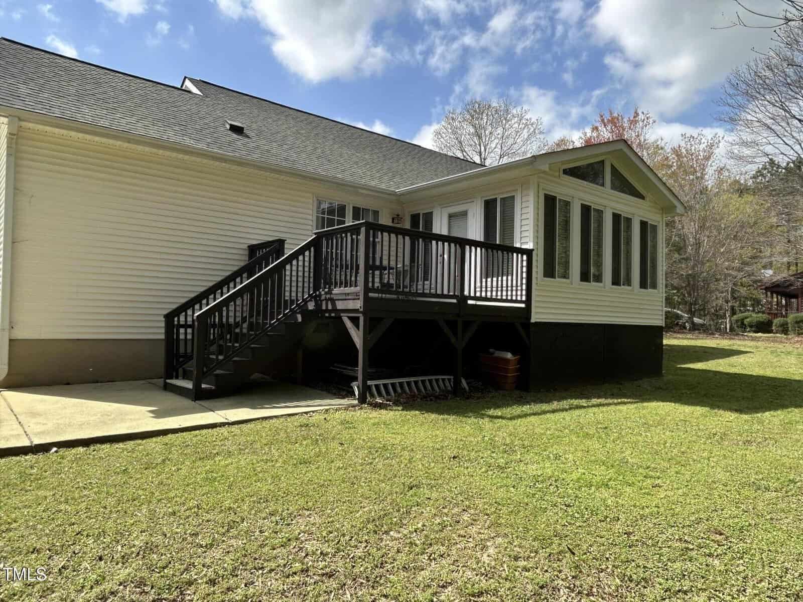 A single-story house with light-colored siding features a small raised deck with black railings, stairs leading to a concrete patio, and an attached sunroom with large windows, surrounded by a grassy yard.