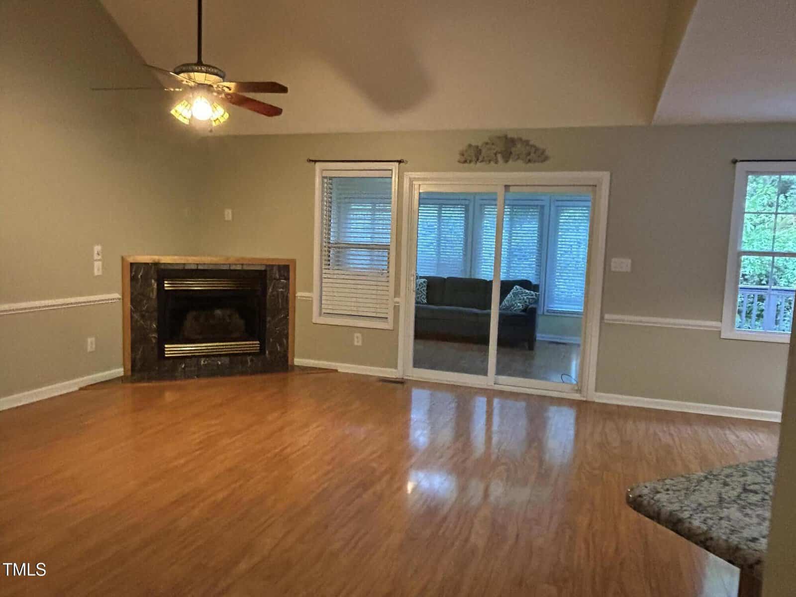 Empty living room with hardwood floors, a ceiling fan with lights, a fireplace in the corner, and sliding glass doors leading to a sunroom with windows. An adjacent window and part of a granite countertop are also visible.