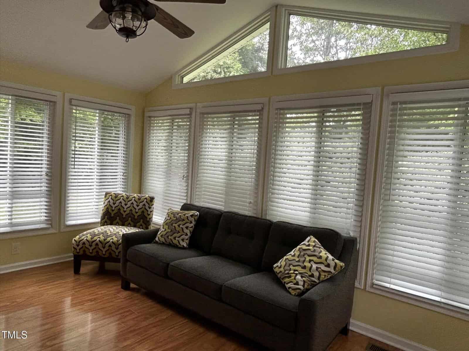 A sunroom with large windows, white blinds, yellow walls, wood flooring, a dark gray sofa with zigzag pillows, a matching chair, and a ceiling fan with a light.