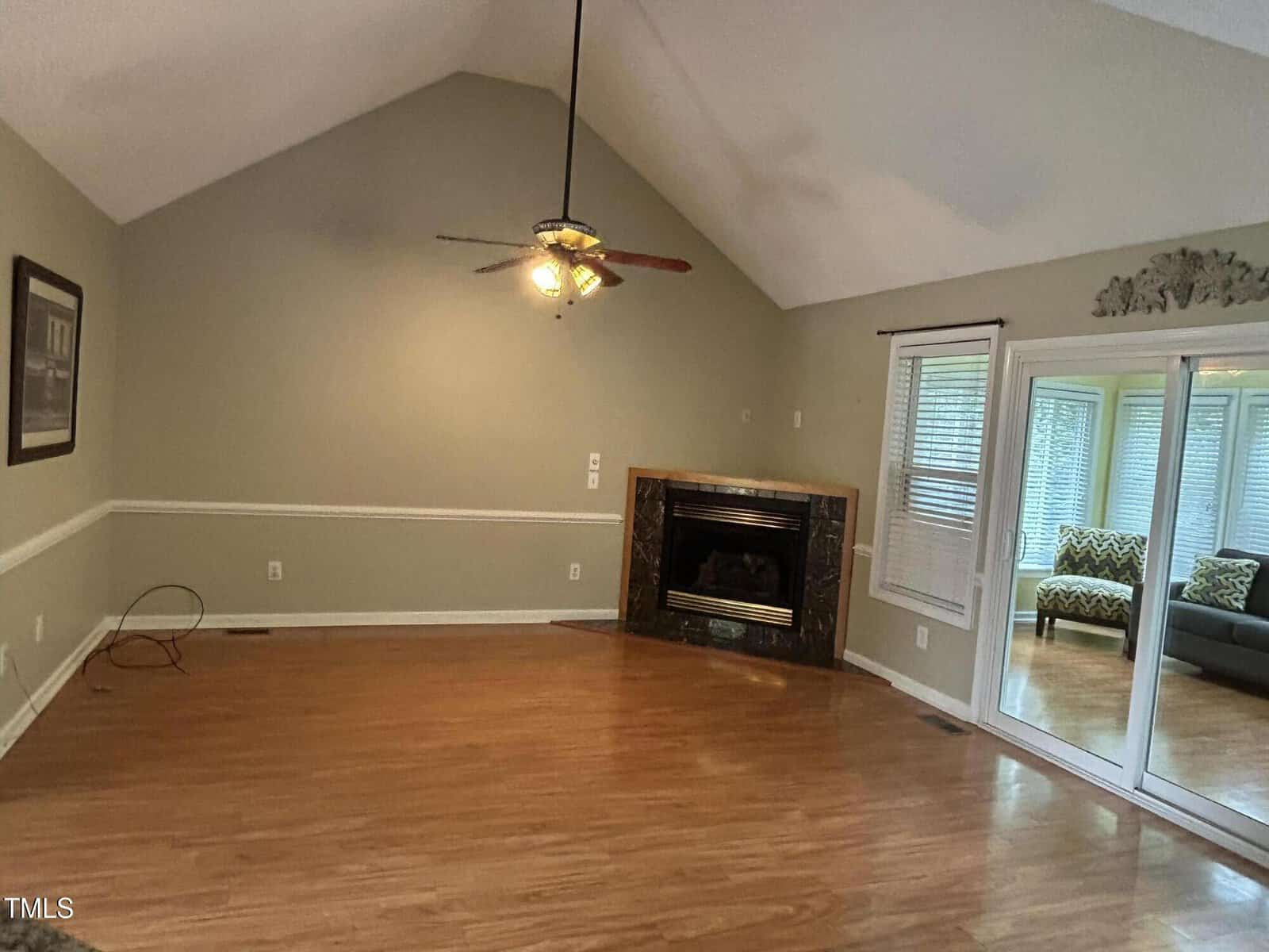Empty living room with wood flooring, a vaulted ceiling, ceiling fan with lights, corner fireplace, light green walls, and a large glass sliding door leading to a sunroom with windows and a patterned chair.