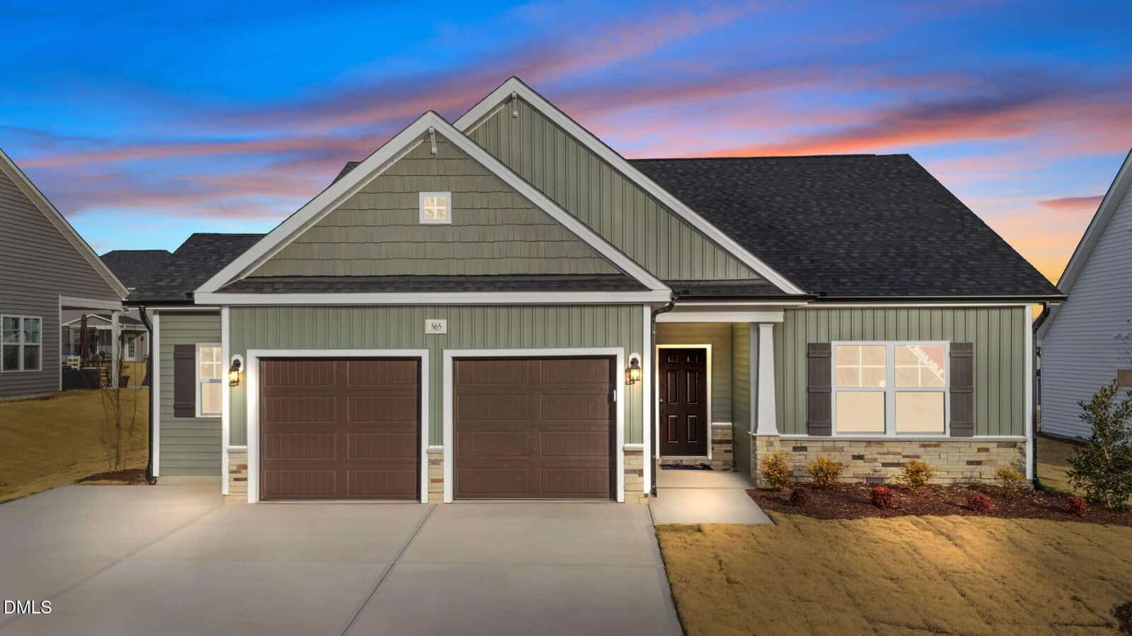 A modern single-story house at 565 Husketh Road, Youngsville, features green siding, brick accents, and two brown garage doors. Exterior lights illuminate the home at dusk under a sky painted with pink, orange, and blue hues. Bushes and mulch line the walkway.
