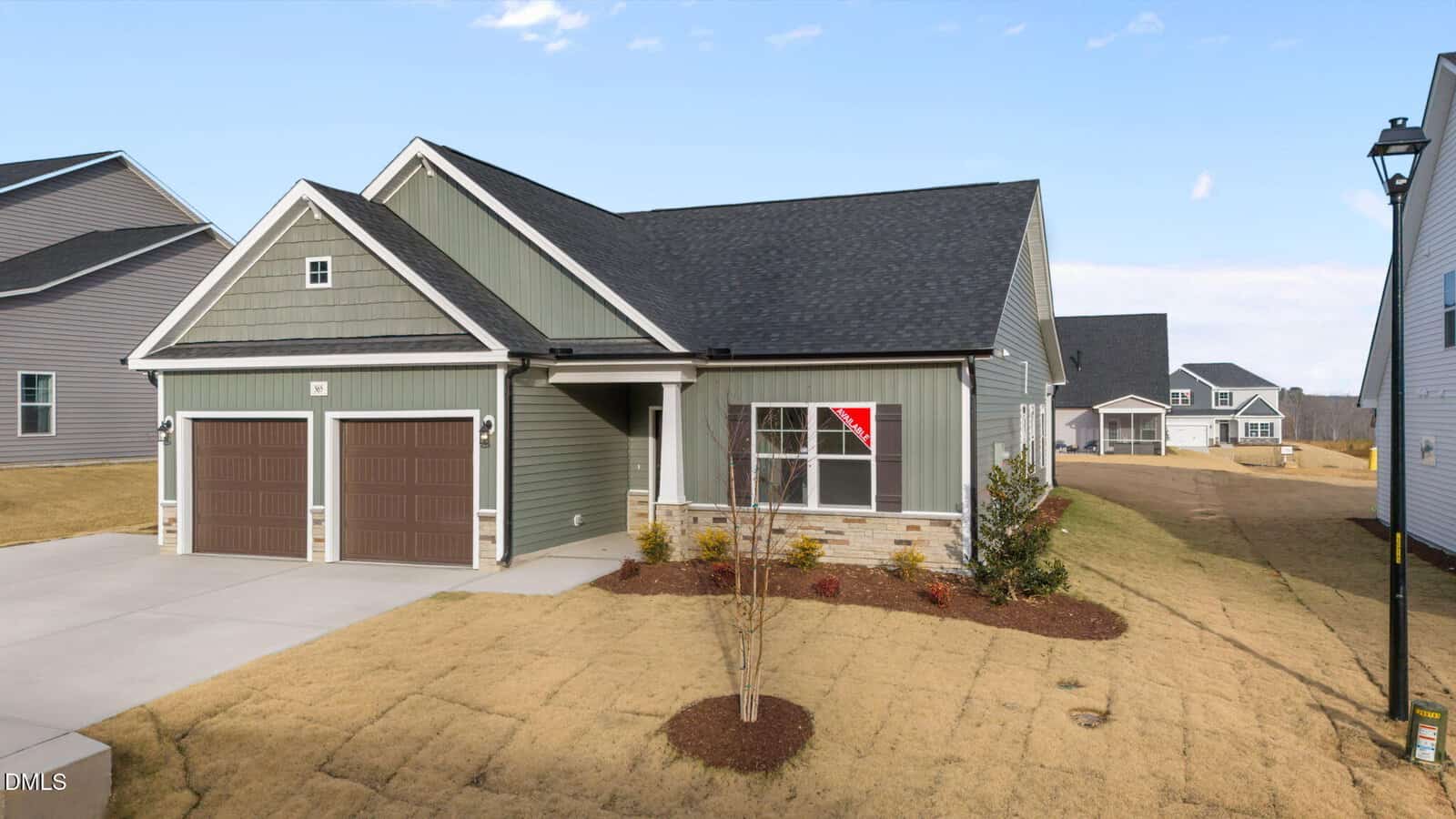 A modern single-story house with green siding, brown double garage doors, and a black shingle roof. The lawn is newly seeded, and a small tree and shrubs are planted near the entrance. A for-sale sign is visible in the front window.