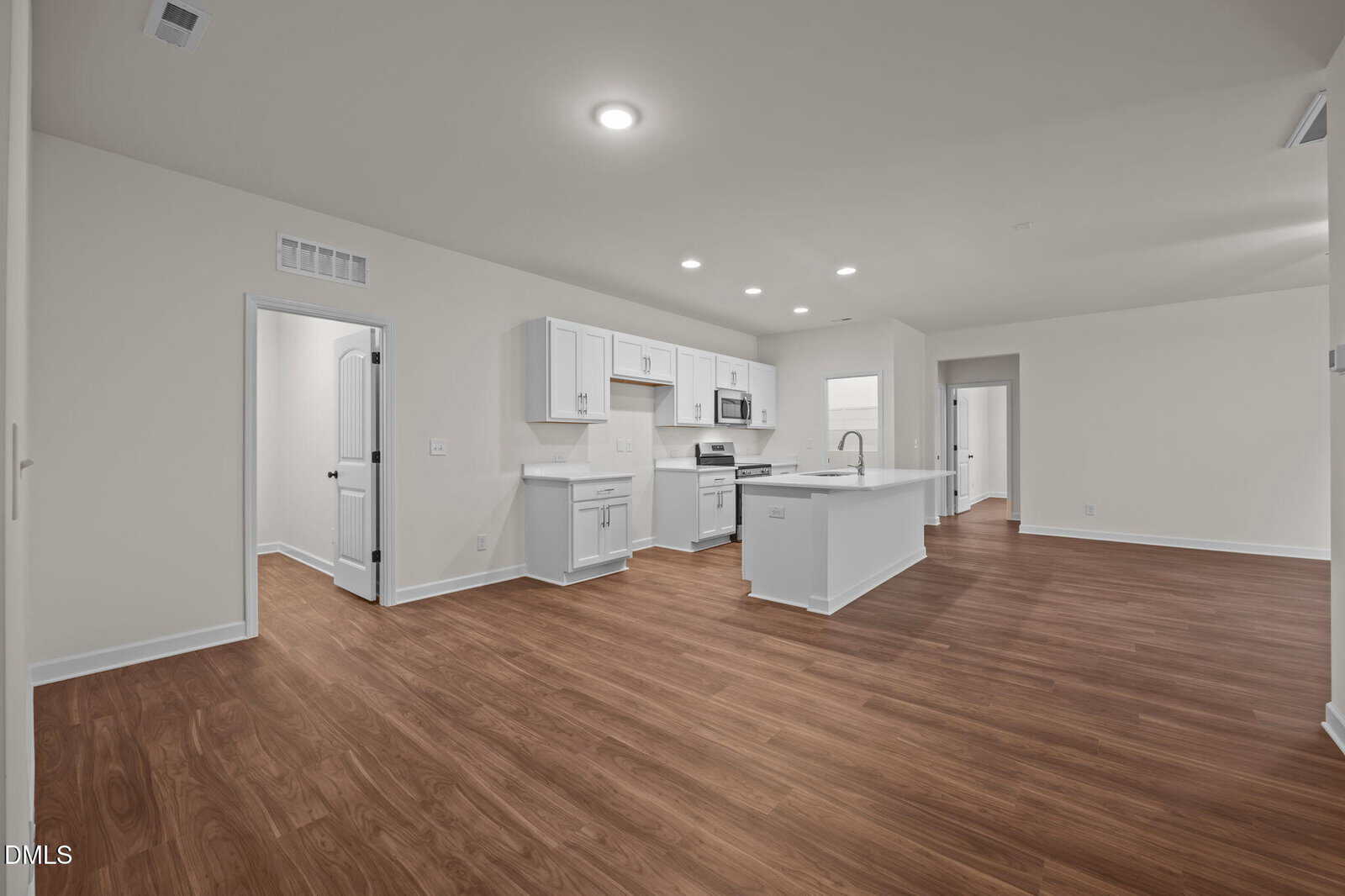Spacious modern kitchen and living area with wood floors, white cabinets, built-in appliances, and recessed lighting. Doorways lead to additional rooms; overall space is clean and unfurnished.