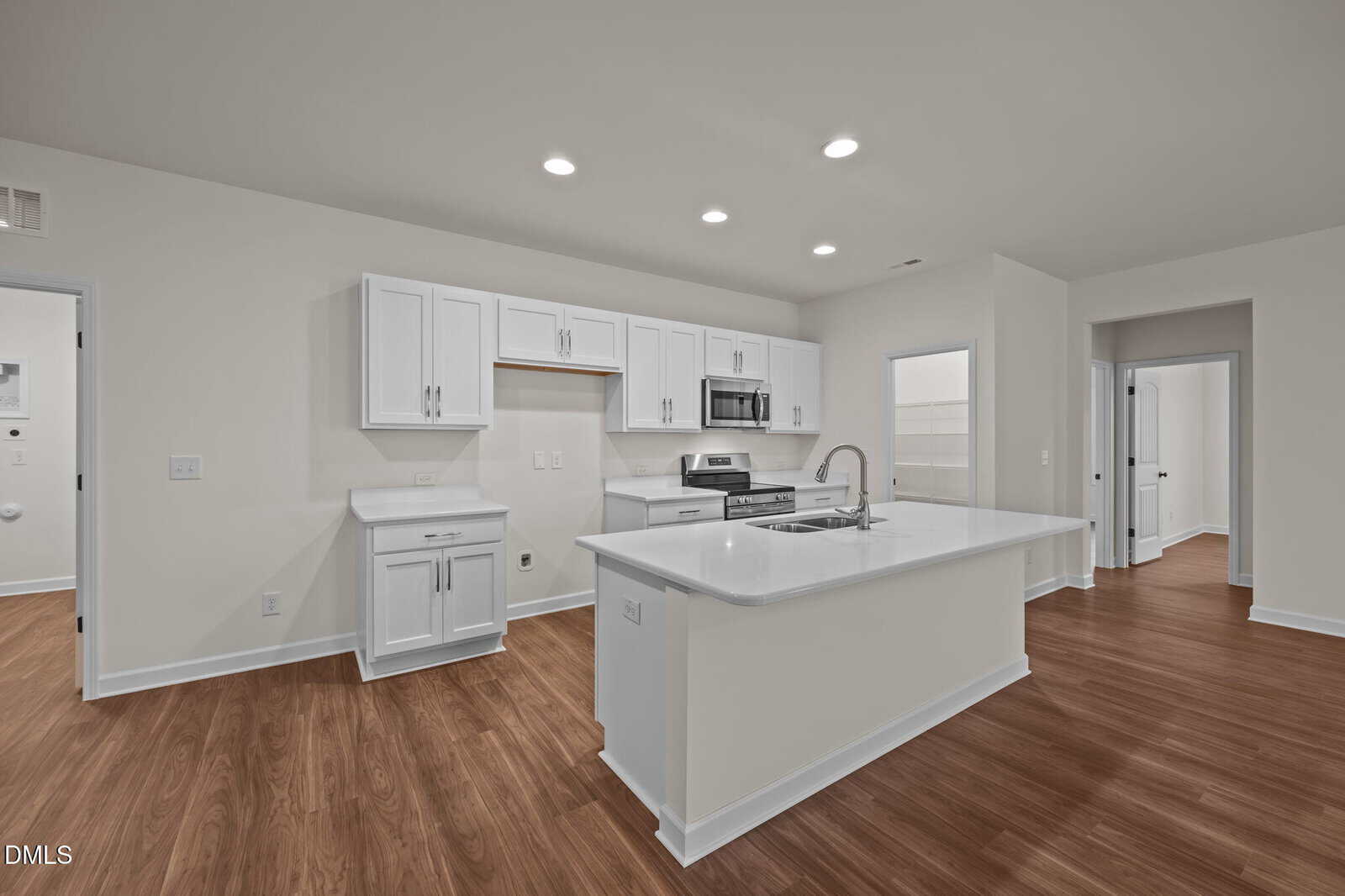 Modern kitchen with white cabinets, stainless steel appliances, and a large island with a sink. The room features wood-style flooring, recessed lighting, and white walls, with doorways leading to adjacent rooms.