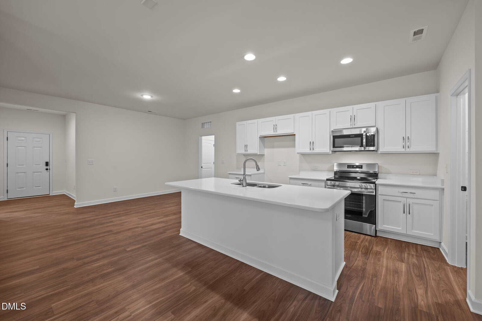 Modern kitchen with white cabinets, stainless steel appliances, and a large island with a sink. Open floor plan with wood-style flooring and recessed lighting, leading to a hallway and front door.