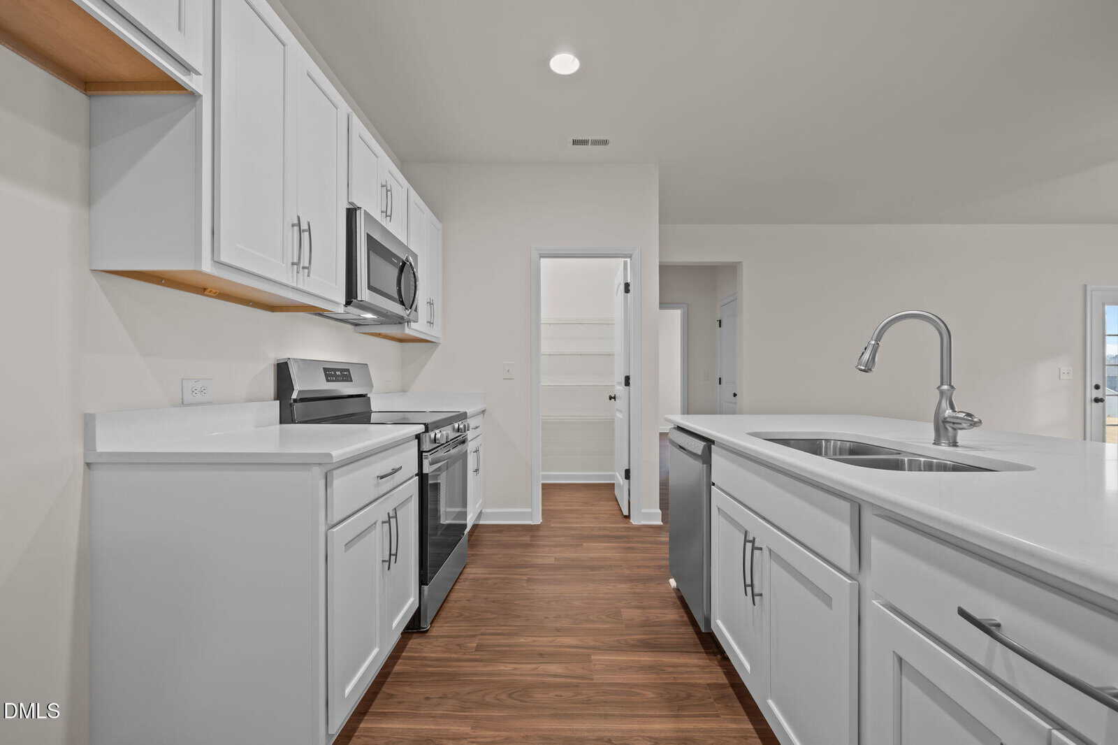 Modern kitchen with white cabinets, stainless steel appliances, and a double sink in an island. The floor is wood, and there’s a door leading to a pantry or storage area in the back. Room is well-lit and spacious.