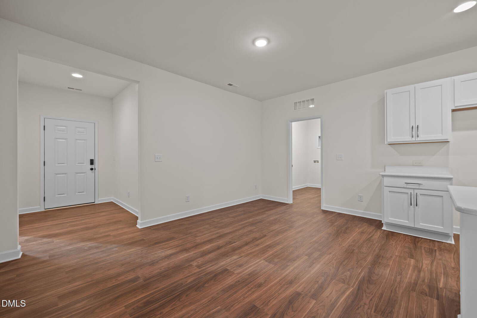 A bright, empty room with white walls, recessed lighting, and wood-style flooring. The space includes white cabinets, a countertop, and doorways leading to other rooms, as well as a white entrance door.