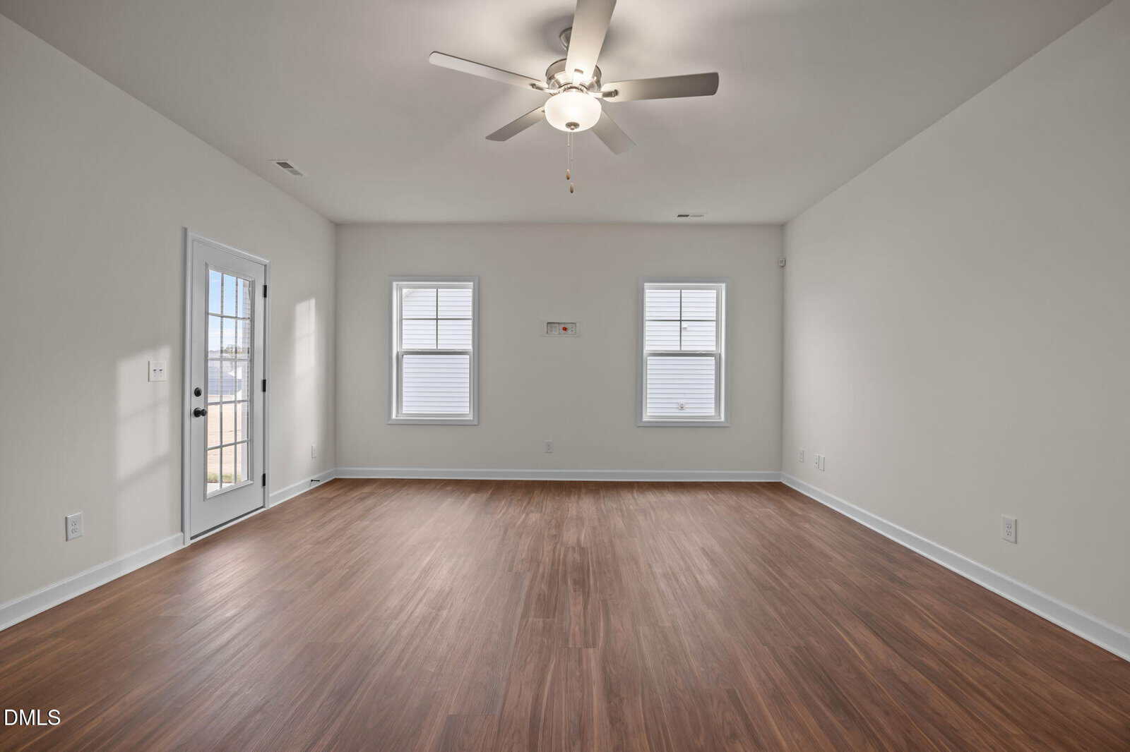 Empty room with white walls, two windows, a ceiling fan, wood-style flooring, and a glass door leading outside. The room appears bright and spacious with natural light coming in through the windows.