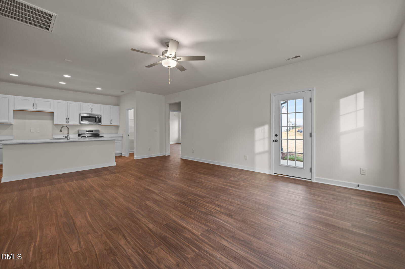 Spacious, empty living area with wood flooring, white walls, ceiling fan, and a door with glass panes. In the background, an open kitchen features white cabinets, stainless steel appliances, and recessed lighting.