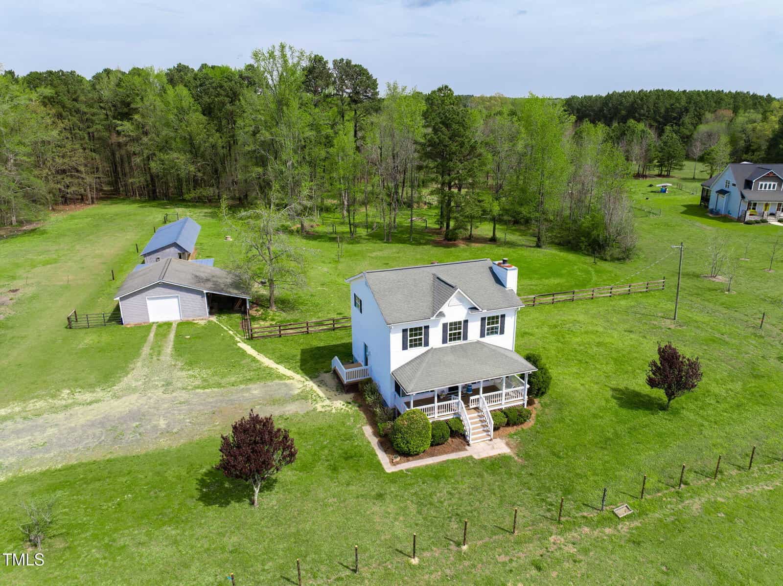 Aerial view of a two-story white house on 2651 Bennett Road, Creedmoor, with a covered front porch, surrounded by lush green grass and trees. A detached garage and shed are nearby. A wooden fence encloses the property, while neighboring houses dot the distant landscape.