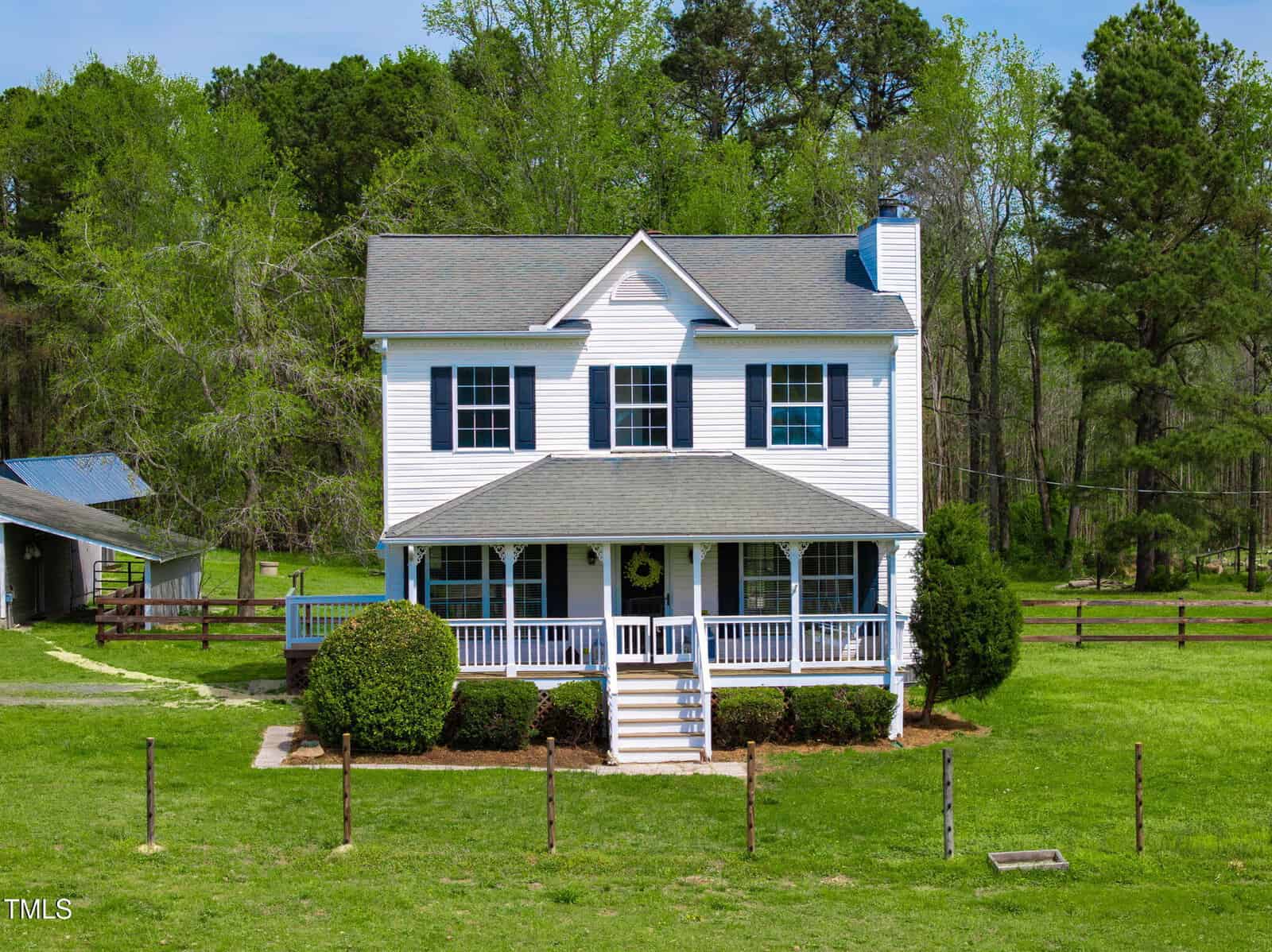 A two-story, white house with a front porch and steps leading to a lawn. Surrounded by trees, a fence, and a small outbuilding. The roof is gray, and there are shrubs in front of the porch. The sky is clear, and the area is grassy.