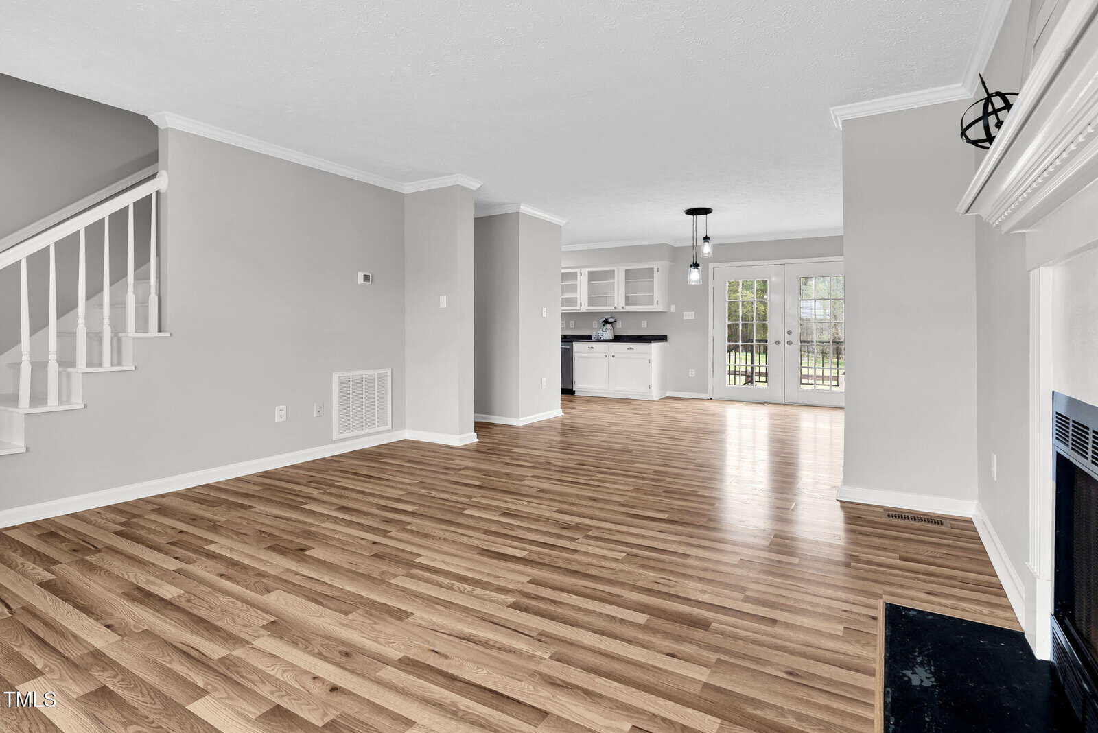 Spacious, unfurnished living area with wood flooring, light gray walls, and a white ceiling. Features include a staircase, fireplace, and open access to the kitchen. Double glass doors in the kitchen lead to an outdoor area.