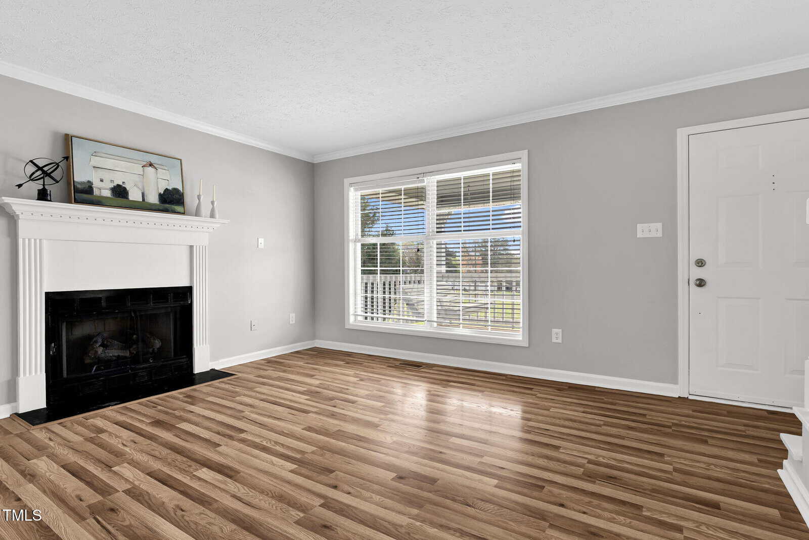 A living room with light gray walls and a white ceiling. It features a white fireplace with a decorative mantel and a large window with blinds. The floor is hardwood. A white door stands on the right side.