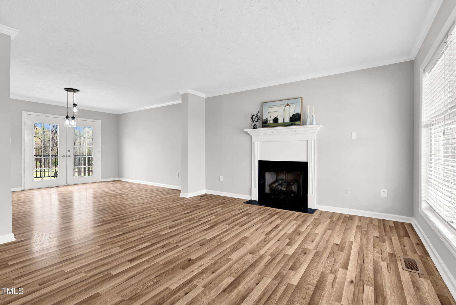 A spacious, empty living room with light wood flooring and light gray walls. A white fireplace is centered on the right wall. Large windows on the right and a hanging light fixture near the back, with double glass doors opening to the outside.