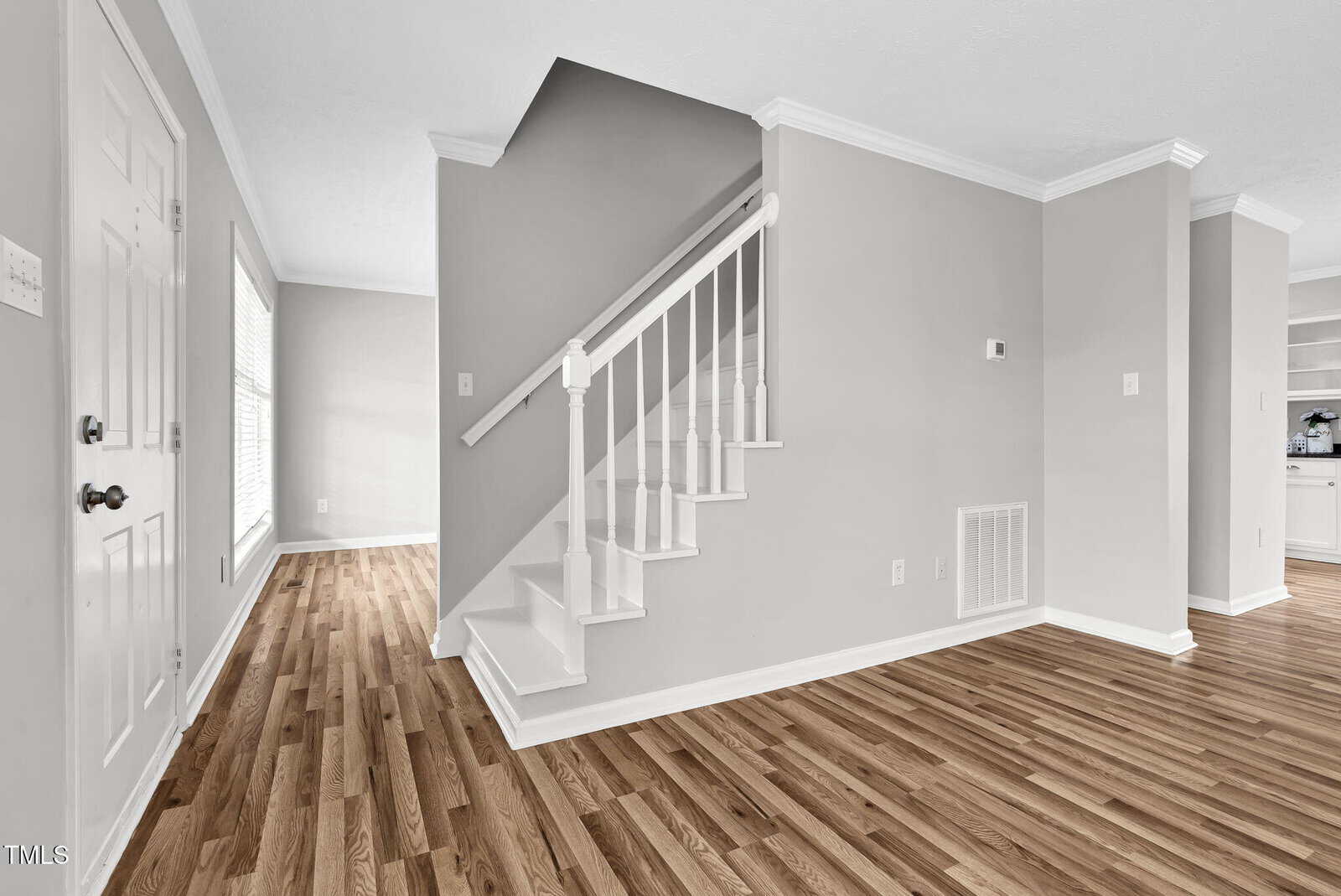 Interior of a house featuring a staircase with white railings and steps, adjacent to a hardwood floor. The walls are painted light gray, and there is natural light coming through a window. A portion of a kitchen is visible to the right.