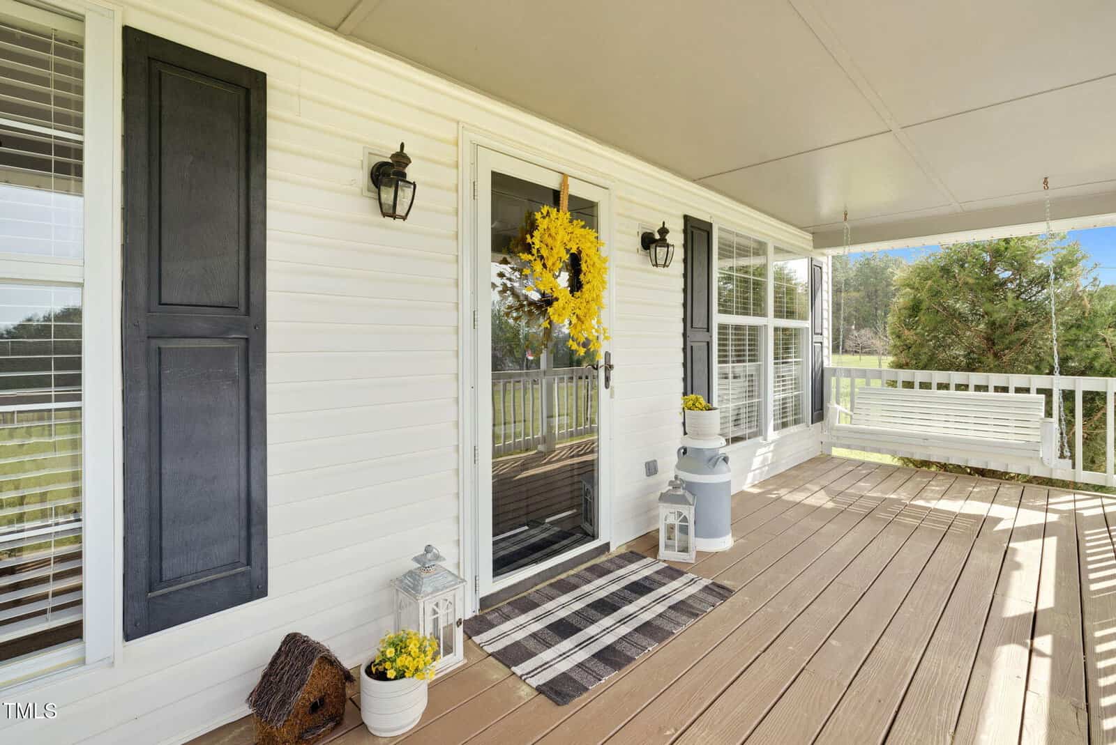 A porch with a wooden floor features a white front door adorned with a yellow floral wreath. There's a black shutter beside the door, a striped doormat, decorative lanterns, and potted plants. A white porch swing is on the right.