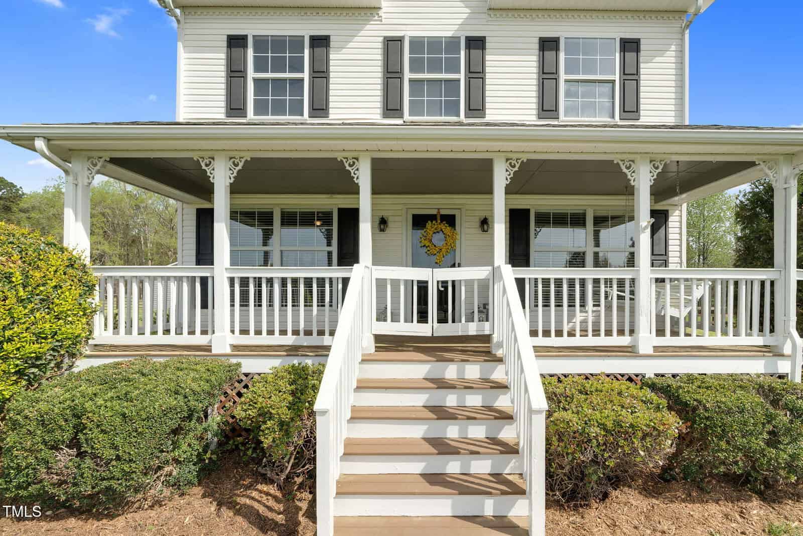 A two-story white house with black shutters and a large front porch. The porch has decorative railings, a wreath on the door, and steps leading down to a garden with shrubs. The sky is clear and blue.