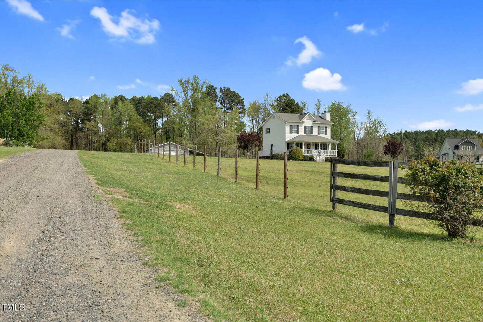 A rural scene features a dirt road on the left and a grassy field with a wooden fence on the right. A two-story white house with a green roof is situated in the background, surrounded by trees under a partly cloudy blue sky.
