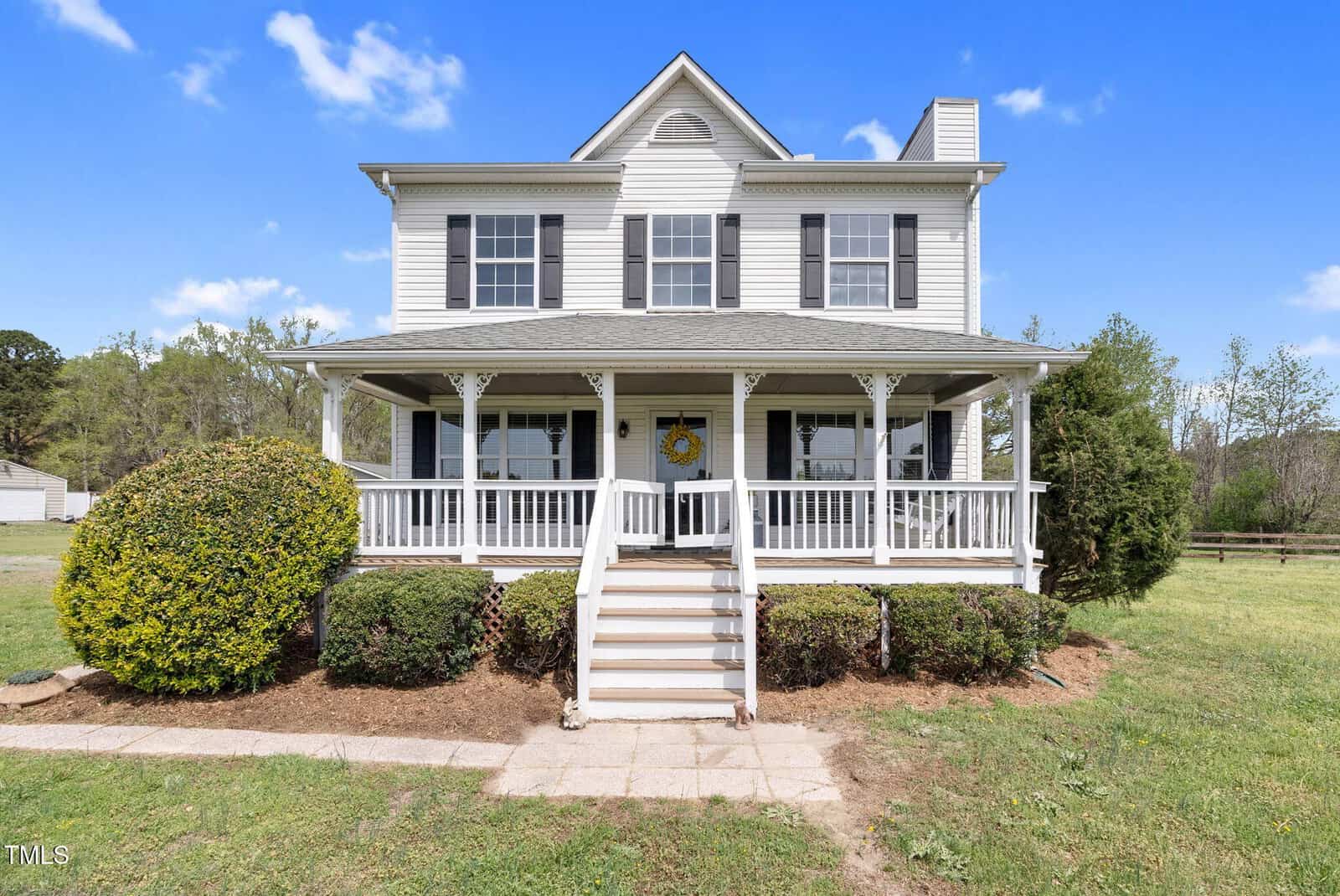 A two-story white house with a wraparound porch and steps leading up to the entrance. It features multiple windows, a chimney, and is surrounded by a well-kept lawn and bushes. The sky is clear with a few clouds.