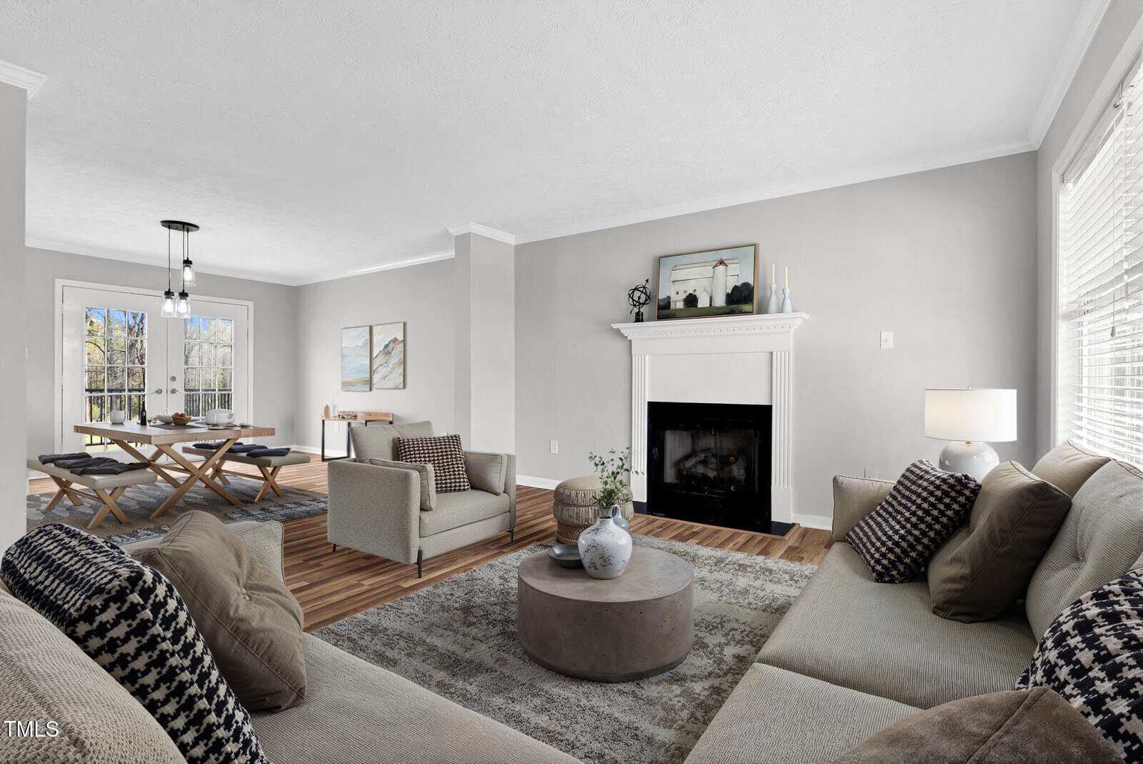 A modern living room with light gray walls, a fireplace with art above it, neutral sofas with patterned pillows, a round coffee table, and large windows. An adjacent dining area with a wooden table is visible in the background.