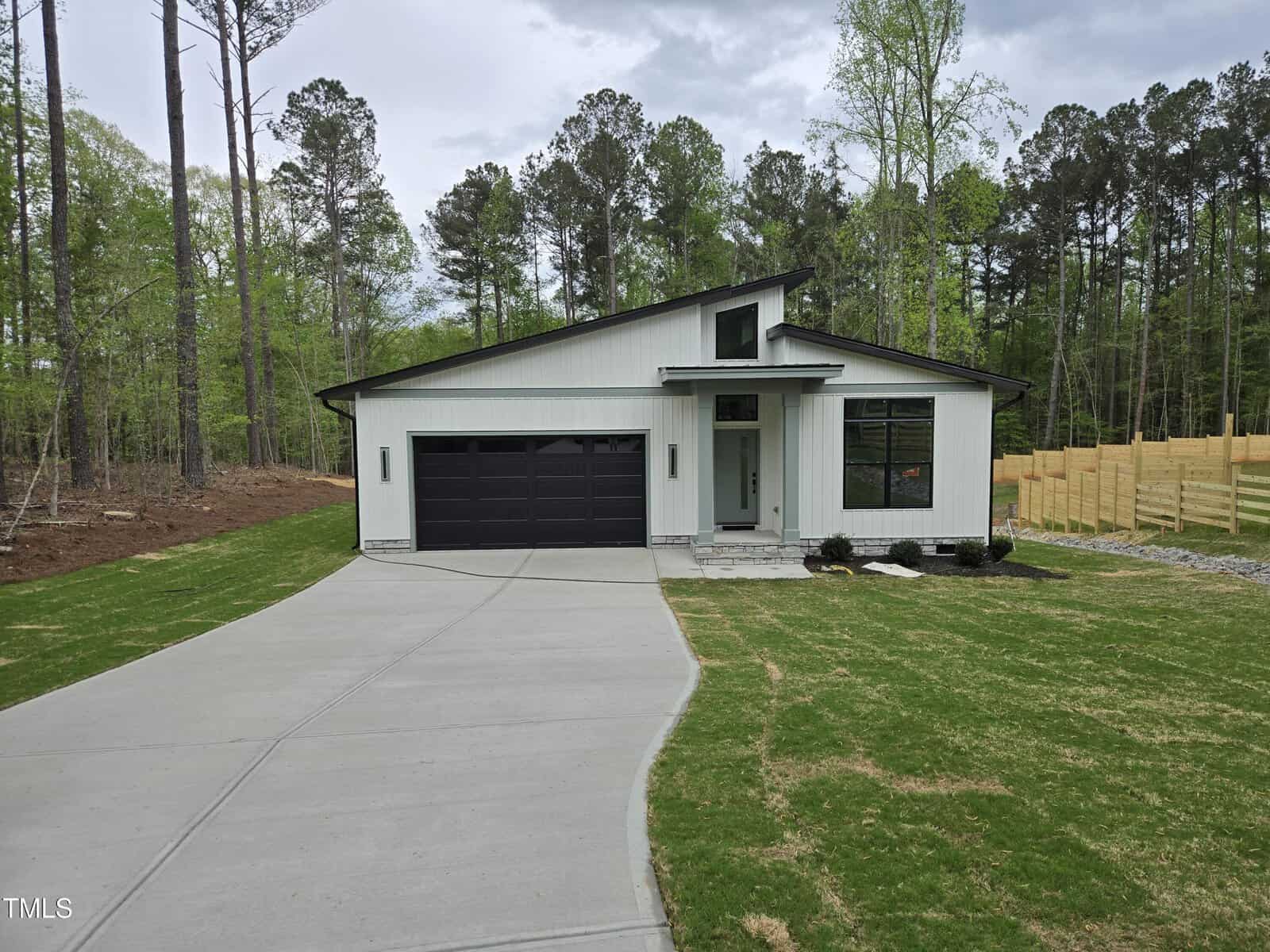 A modern single-story house with white siding, black trim, and a dark garage door. The home has large windows, a sloped roof, and is surrounded by a lawn, trees, and a wooden fence in the background.