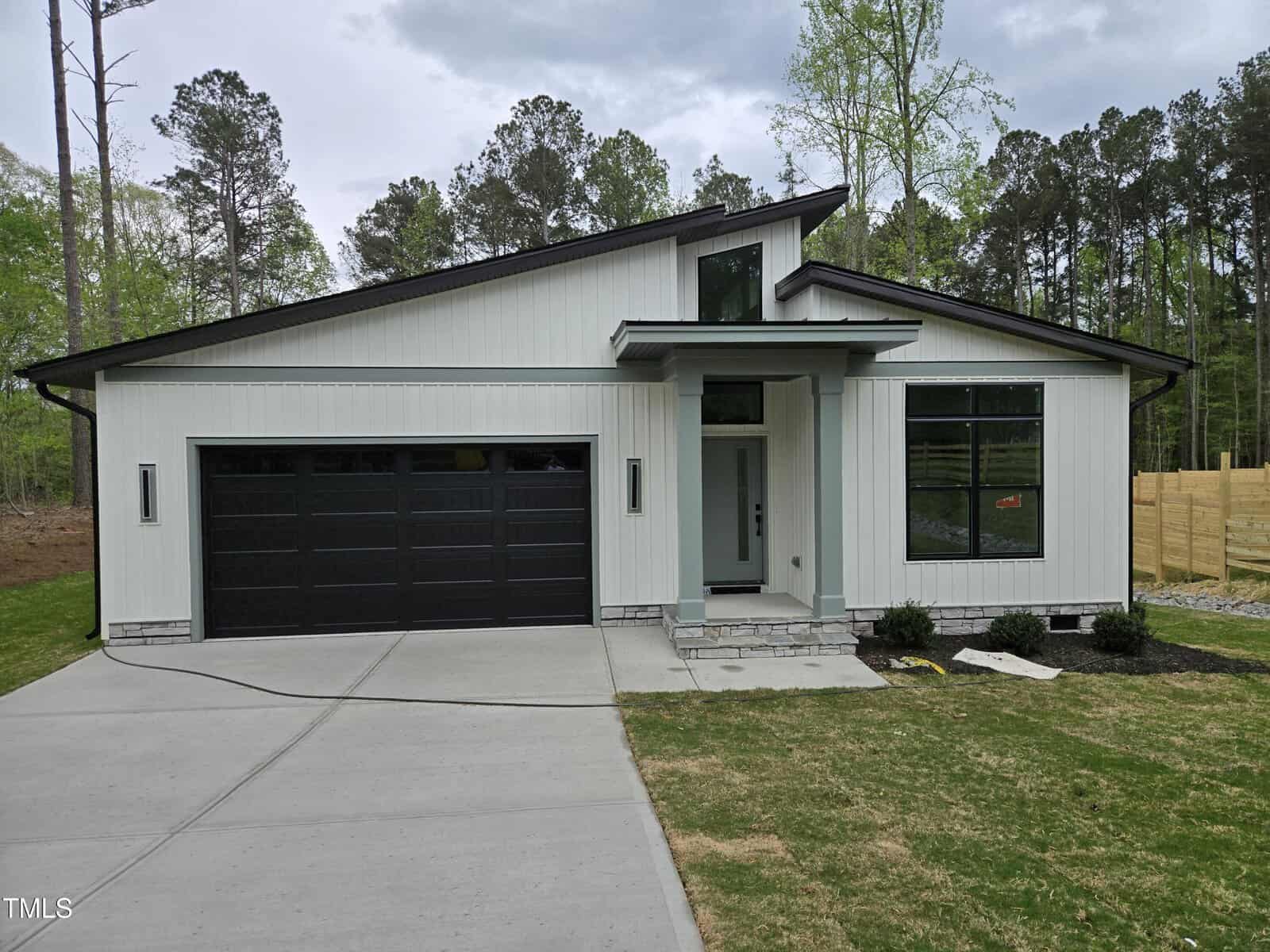 A modern single-story house with white siding, black trim, and a black double garage door. The home has large windows, a small porch, and sits on a concrete driveway with grass and trees in the background.