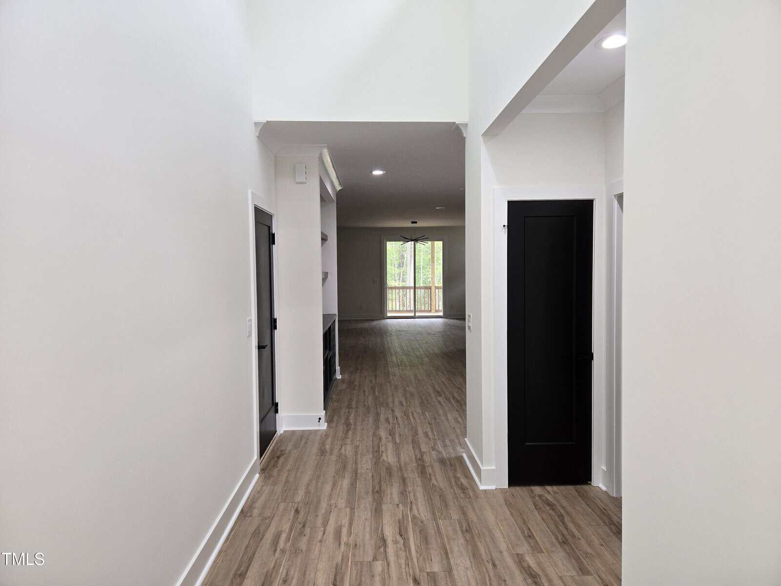 A hallway with light-colored walls and wood flooring leads to a living area with built-in shelves and large windows overlooking a deck and greenery outside. Two black doors are on the right side of the hallway.
