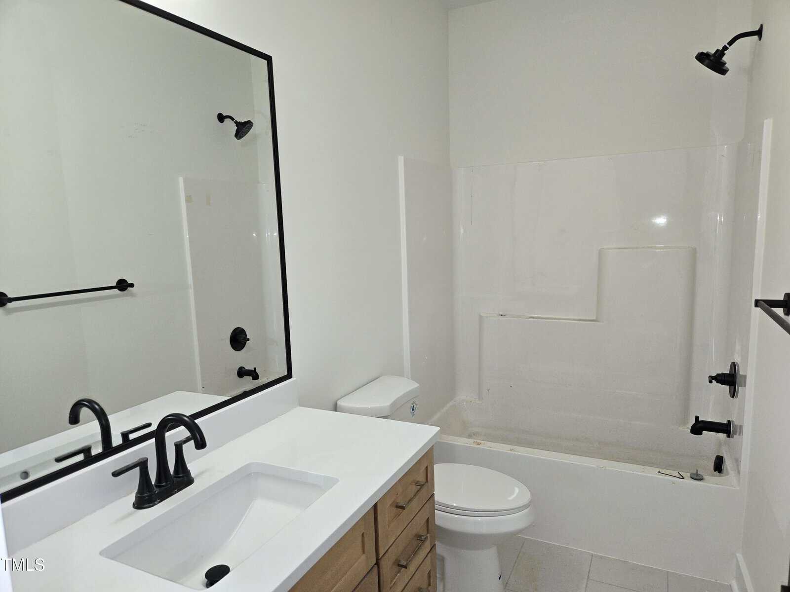 A bathroom with white walls, a white bathtub and shower combination, a white toilet, a wooden vanity with a white countertop, a rectangular mirror, and black fixtures for the faucet, shower, and towel bars.