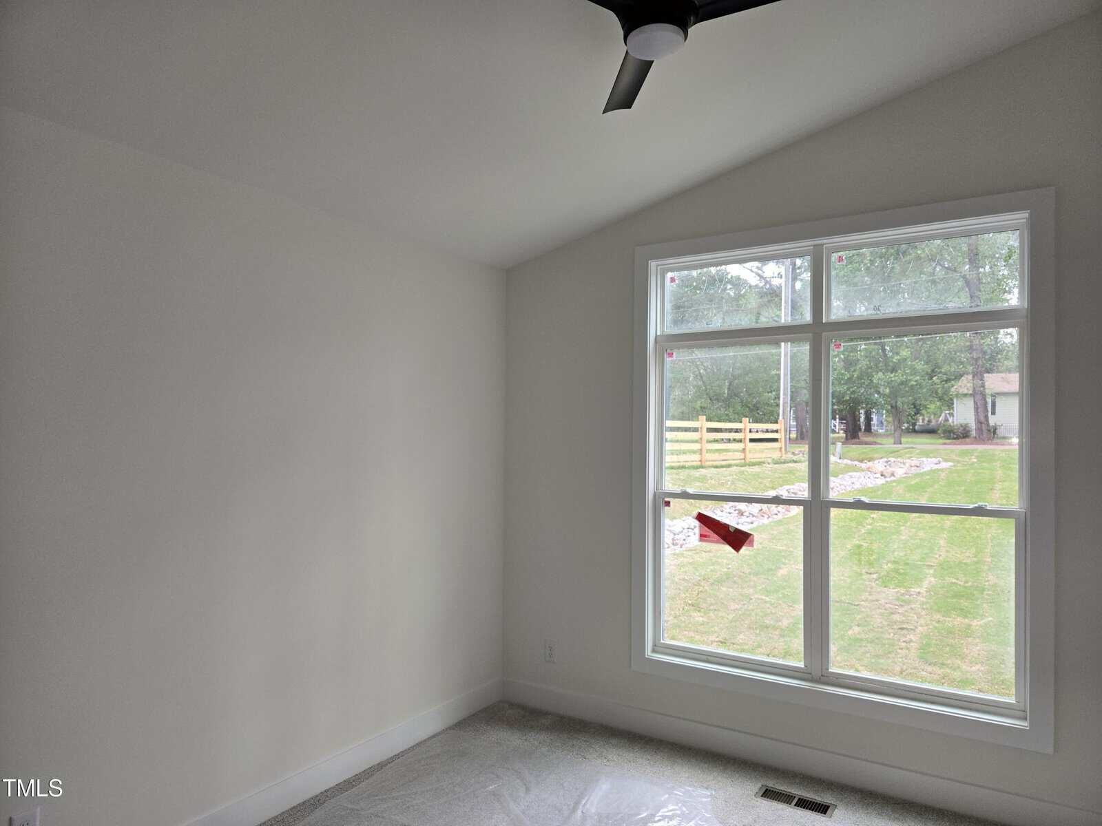 An empty room with white walls, carpeted floor, a ceiling fan, and a large window overlooking a grassy yard with trees and a wooden fence outside.
