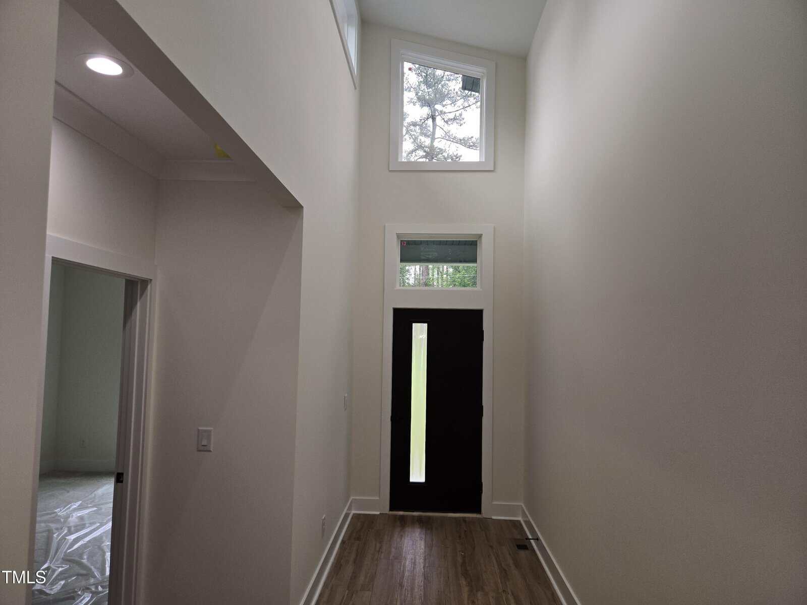 A hallway features a black front door with a tall window above it, letting in natural light. There is a high ceiling, light-colored walls, and wood flooring. An open doorway is visible on the left side.