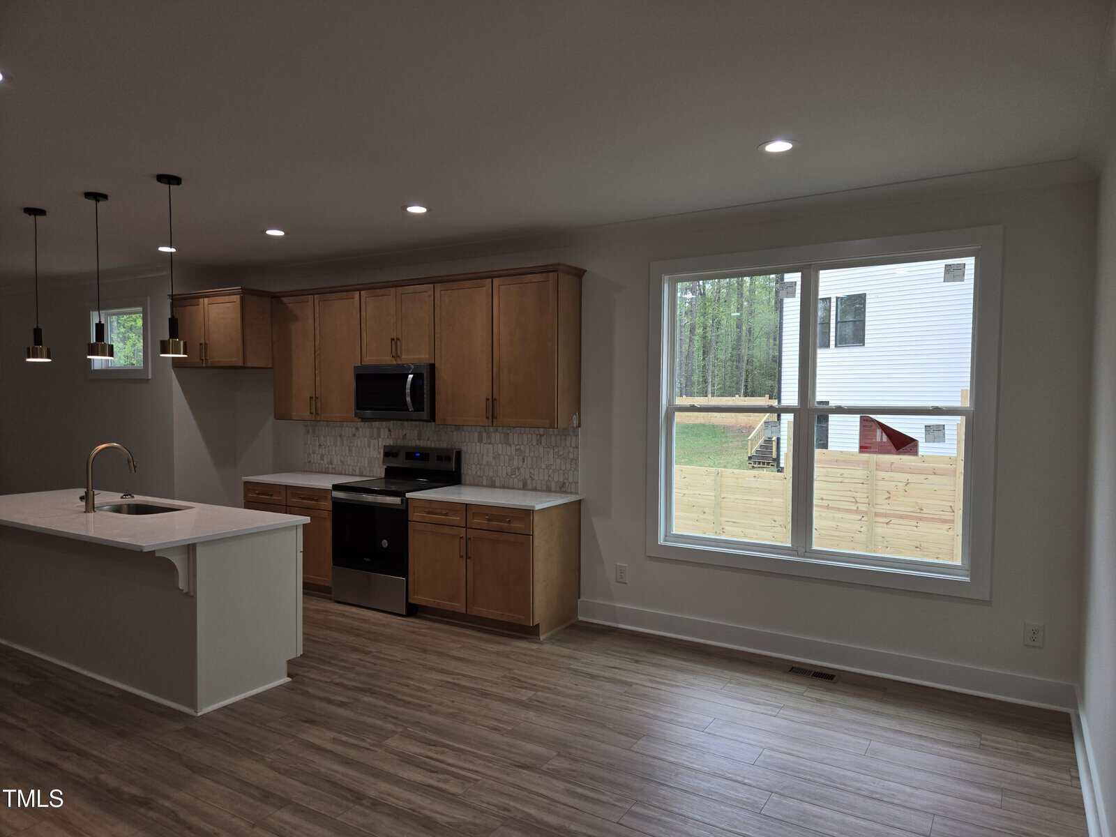 Modern kitchen with light wood cabinets, stainless steel appliances, and an island with a sink; large window overlooks a fenced backyard with trees and a neighboring white house. Wood-look flooring throughout.