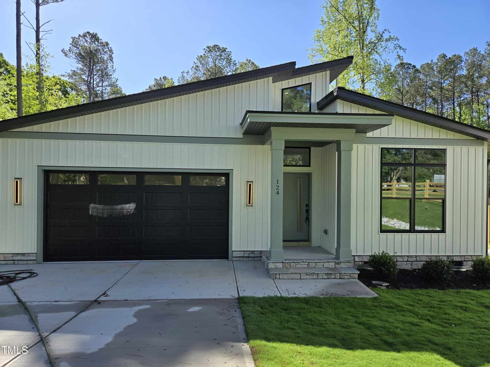A modern single-story house with light-colored vertical siding, a dark two-car garage door, a small covered entryway, rectangular windows, and a neat lawn bordered by shrubs and trees.