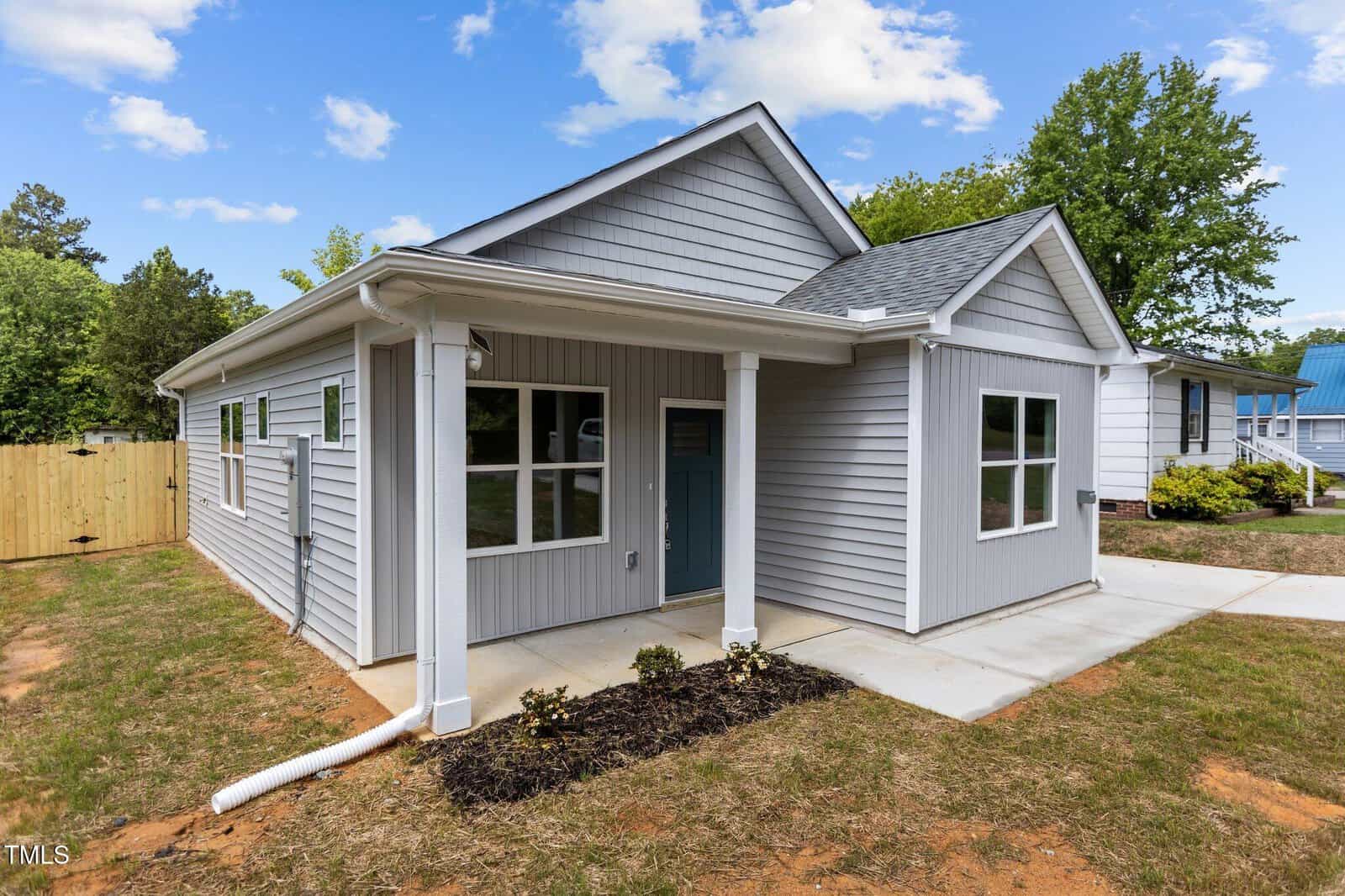 A small, single-story house with gray siding and a covered front porch, set on a lawn with minimal landscaping. The house has white trim, multiple windows, and a driveway on the right side.