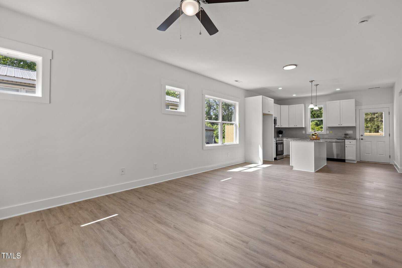 A modern open-concept living area with light wood flooring, white walls, three square windows, ceiling fan, and a view into a white kitchen with stainless steel appliances and an island.
