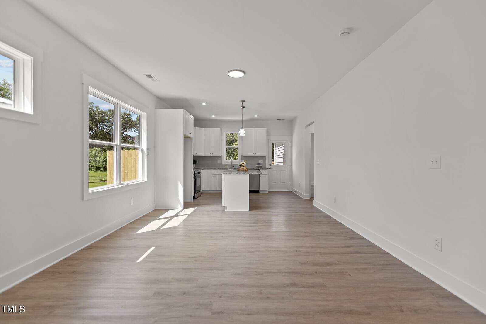 A bright, empty room with light wood floors and white walls leads to a modern kitchen area with white cabinets, an island, and stainless steel appliances. Large windows provide natural light and a view of the outdoors.