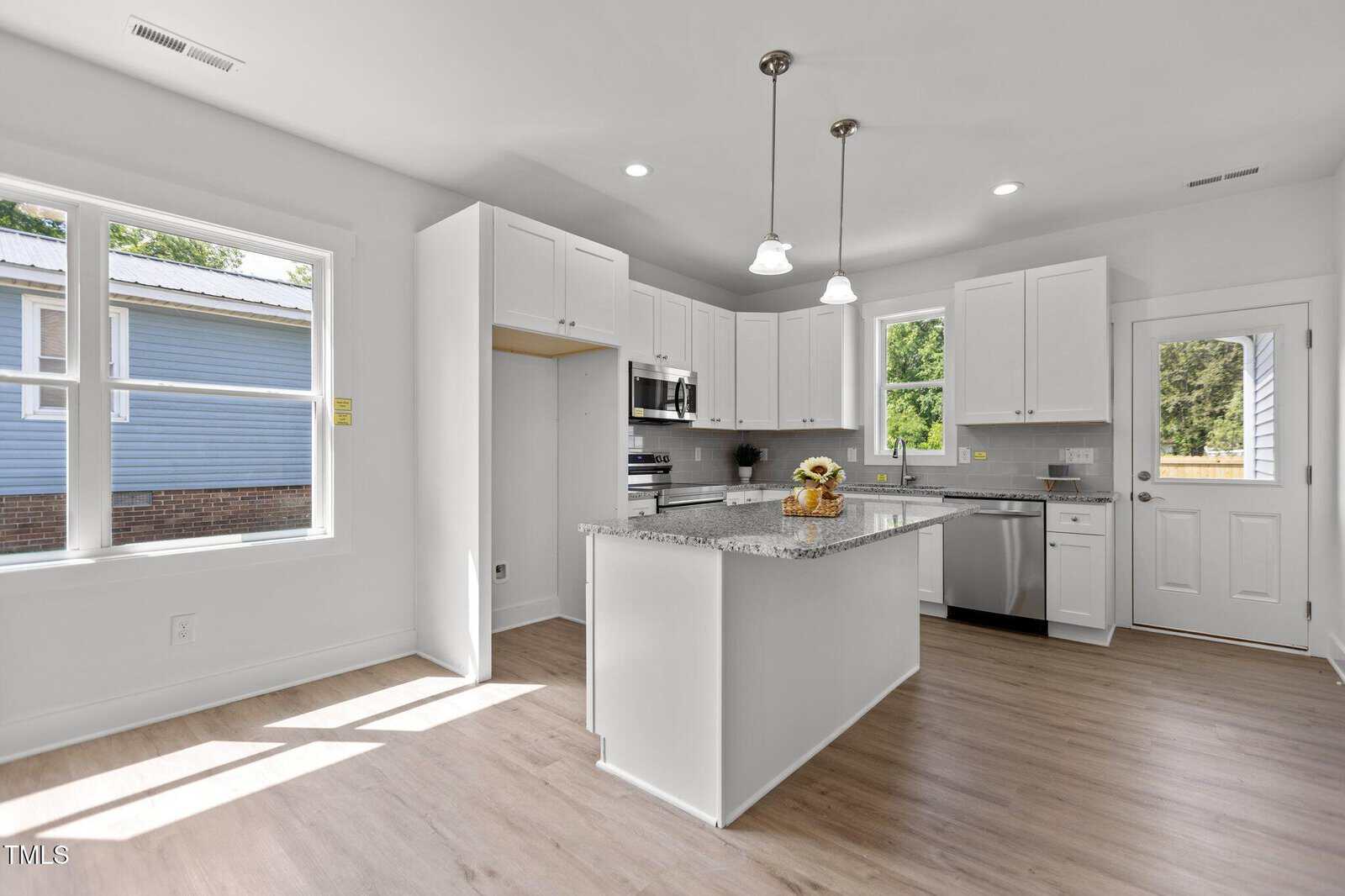 A modern kitchen with white cabinets, stainless steel appliances, a granite island with a vase of sunflowers, light wood floors, large windows, and pendant lighting. There's a door and a window at the back.