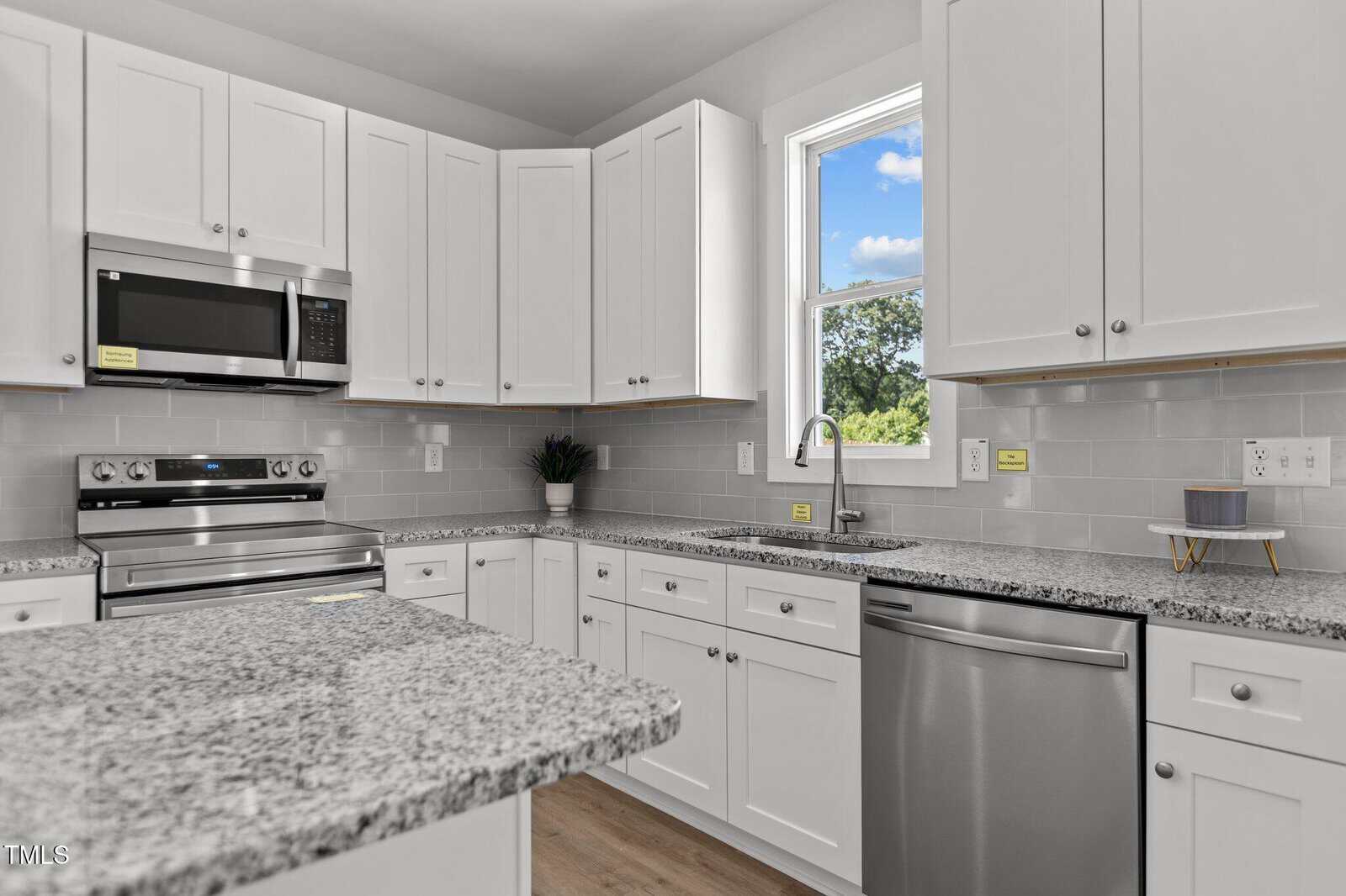 A modern kitchen with white cabinets, stainless steel appliances, gray subway tile backsplash, granite countertops, and a window above the sink letting in natural light.