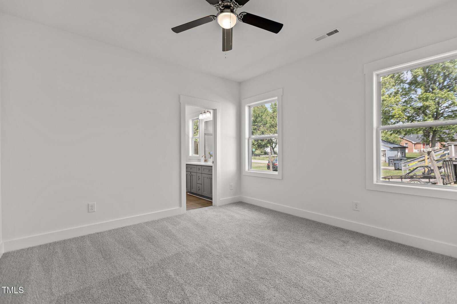 A bright, empty bedroom with light gray carpet, white walls, a ceiling fan with light, two windows, and a doorway leading to a bathroom with a double-sink vanity and outdoor views.