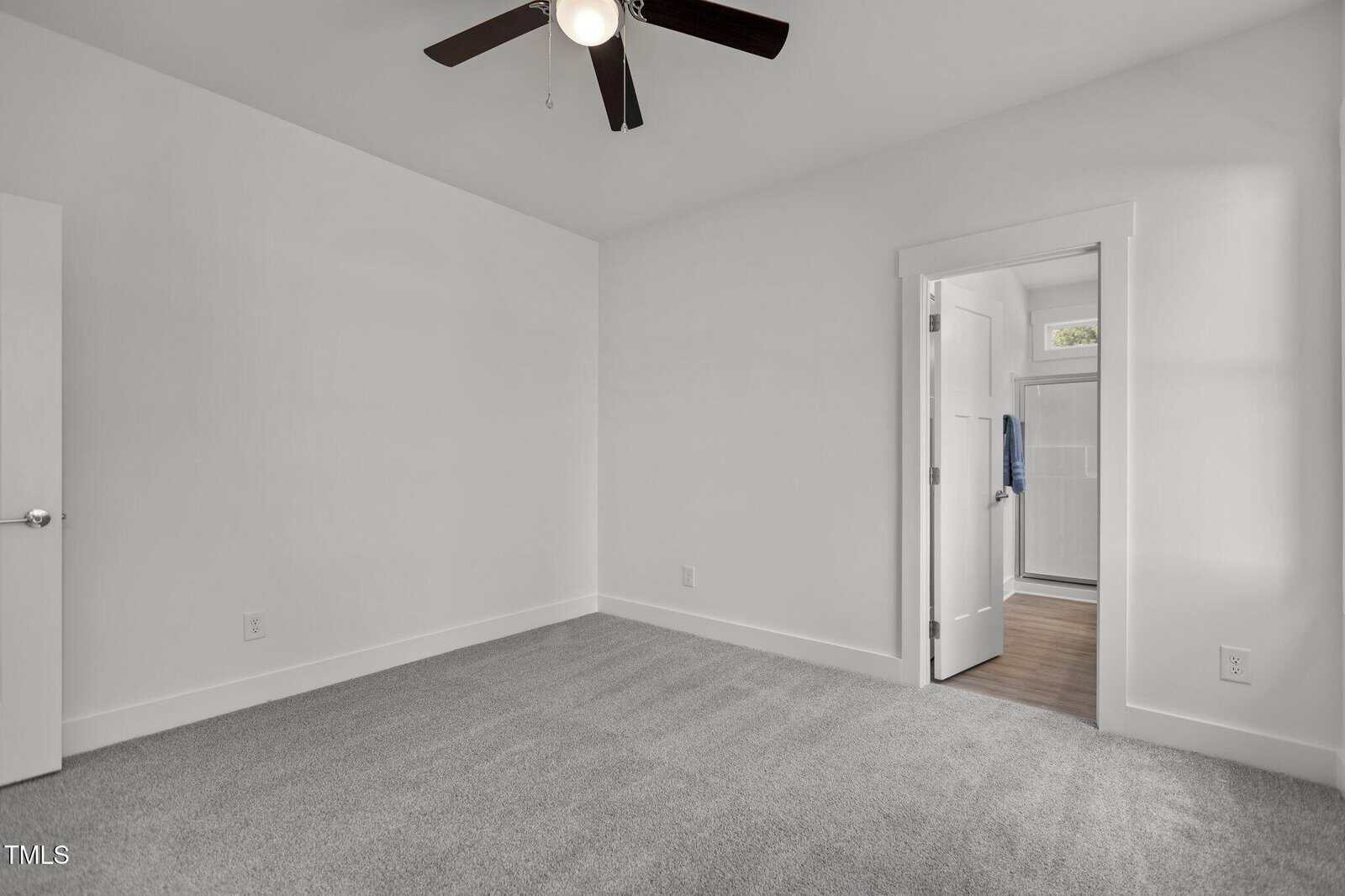 A small, empty room with white walls, gray carpet, a ceiling fan with dark blades, and an open door leading to a bright hallway with wood flooring.