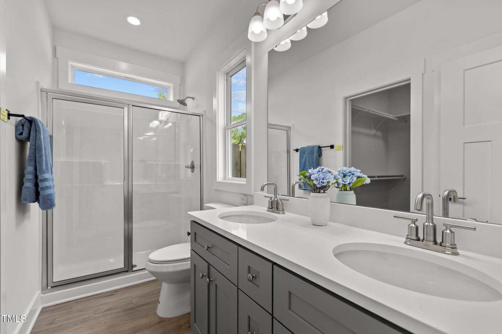 A modern bathroom with a double-sink vanity, gray cabinets, a large mirror, a glass shower enclosure, a toilet, white walls, wood flooring, and a potted flower arrangement on the counter.