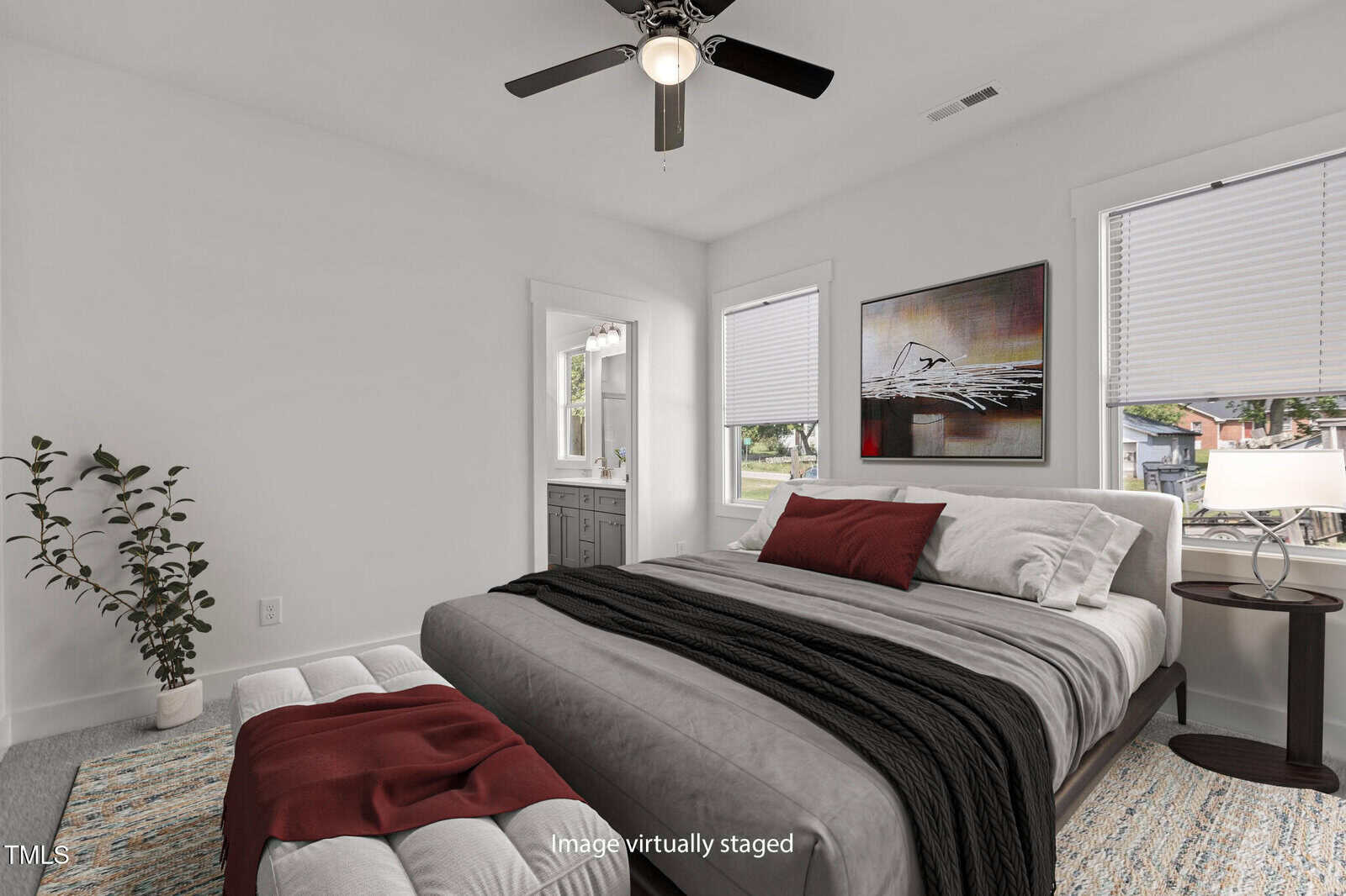 A bedroom with a large bed featuring gray and red bedding, a small side table with a lamp, a framed artwork on the wall, two windows, a ceiling fan, a plant in the corner, and an open doorway to a bathroom.