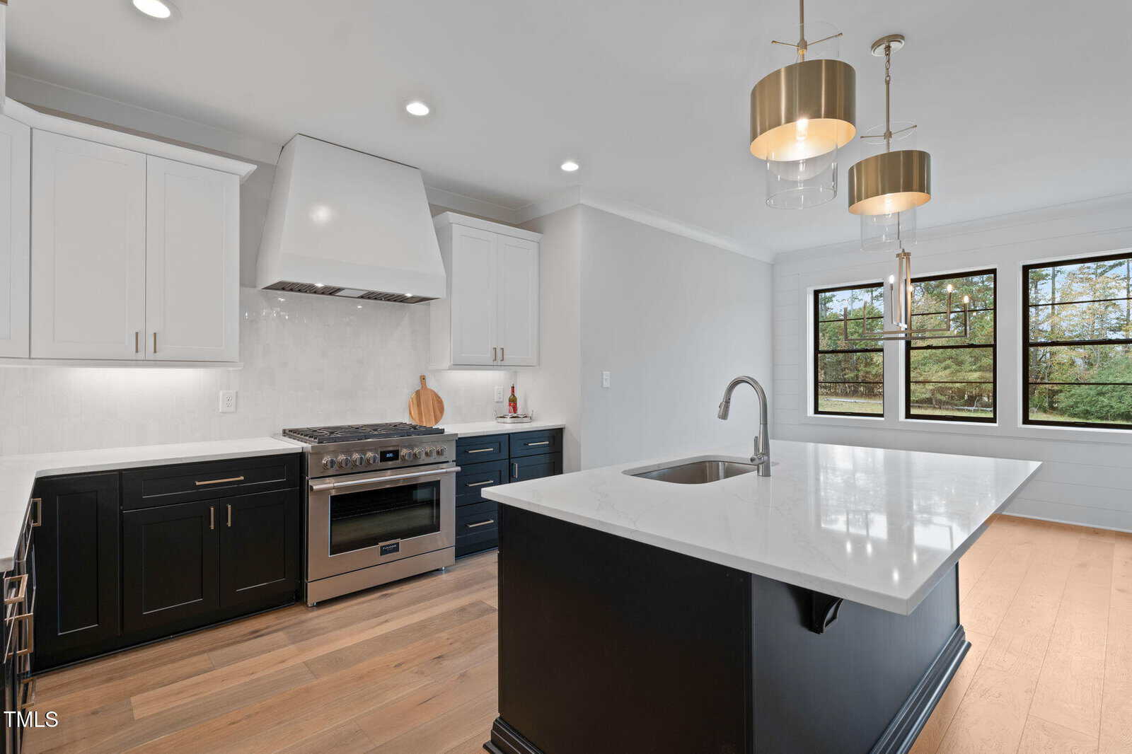 Modern kitchen with white upper cabinets, black lower cabinets and island, stainless steel stove and hood, pendant lights over the island, and large windows showing trees outside. Light wood floor throughout.
