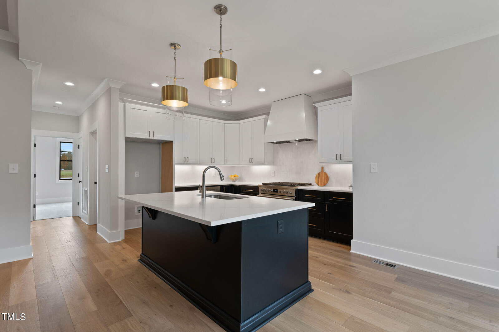 Modern kitchen with white upper cabinets, black lower cabinets and island, white countertops, stainless steel appliances, wood flooring, gold pendant lights, and recessed lighting. A doorway and large window are visible in the background.