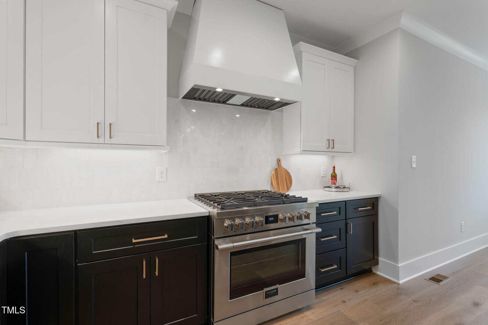 A modern kitchen features a stainless steel gas stove with a large range hood, white upper cabinets, black lower cabinets, and a white backsplash. A wooden cutting board and a bottle of oil sit on the right countertop.