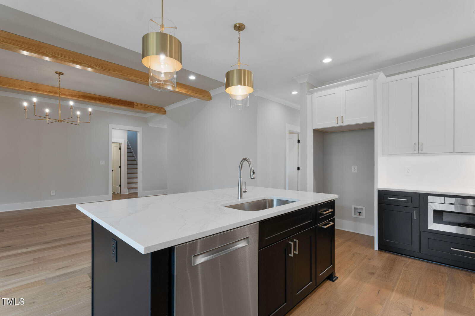 Modern kitchen with a large marble island, stainless steel appliances, black lower cabinets, white upper cabinets, gold pendant lights, wood floor, and exposed wooden ceiling beams in an open floor plan.