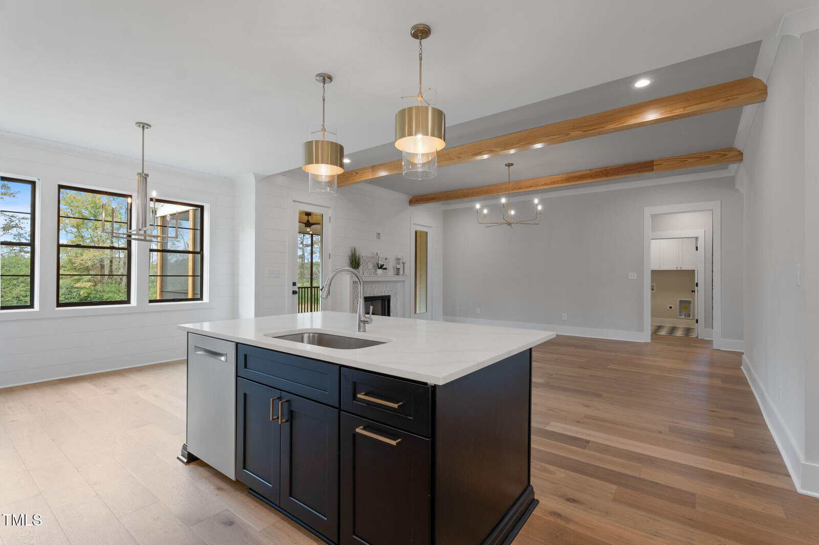 Modern open kitchen and living area with a dark island, white countertops, wood floors, exposed ceiling beams, pendant lights, large windows, and a visible doorway leading to another room.