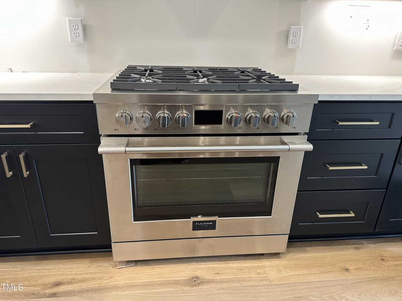A stainless steel gas range with six burners, control knobs, and an oven sits between dark blue cabinets with brass handles on a wood floor. White wall and countertop are visible in the background.