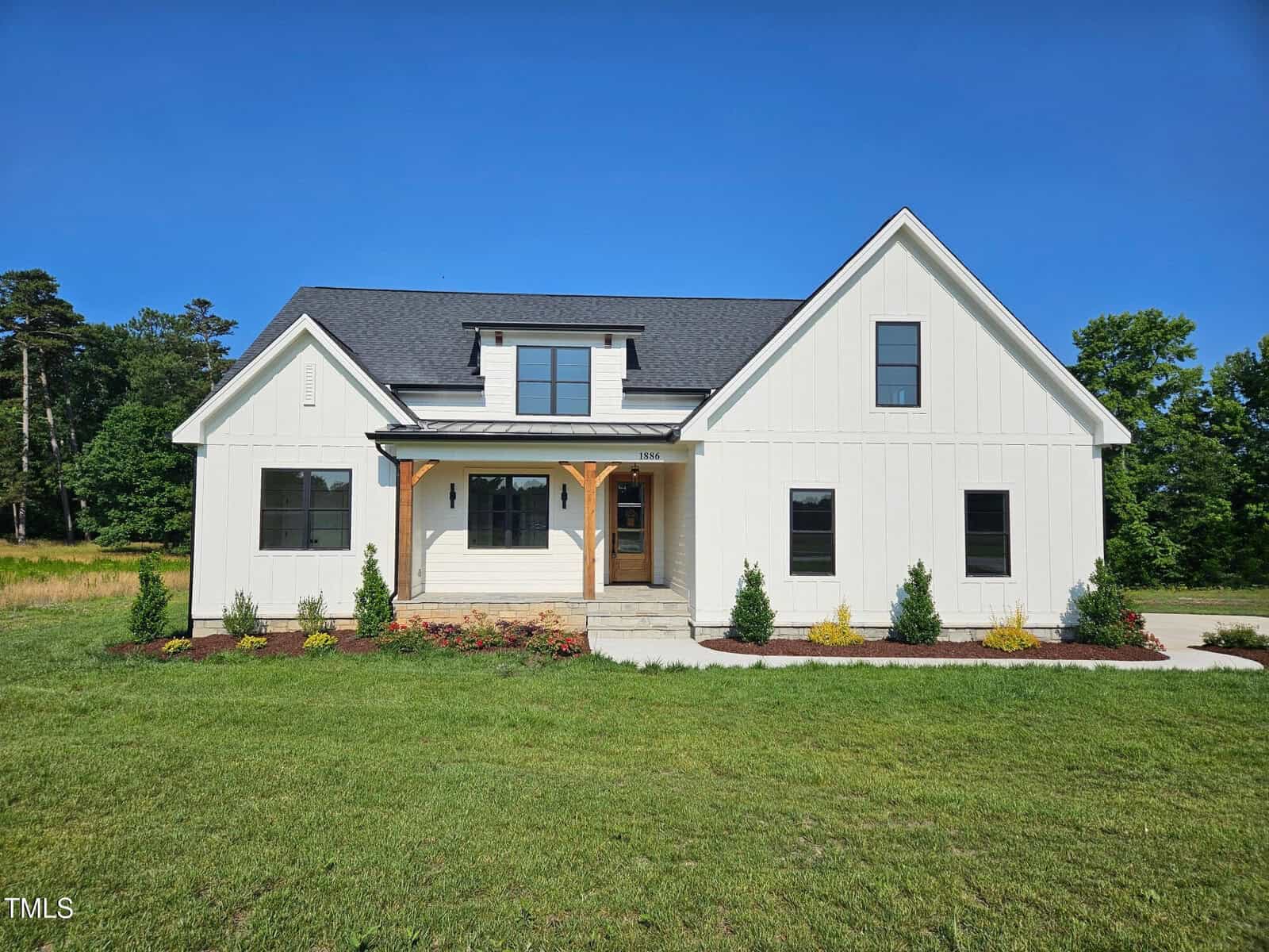 A modern white farmhouse with a dark roof, wooden accent posts on the porch, large front windows, and a neatly landscaped yard, set against a clear blue sky and green grass.