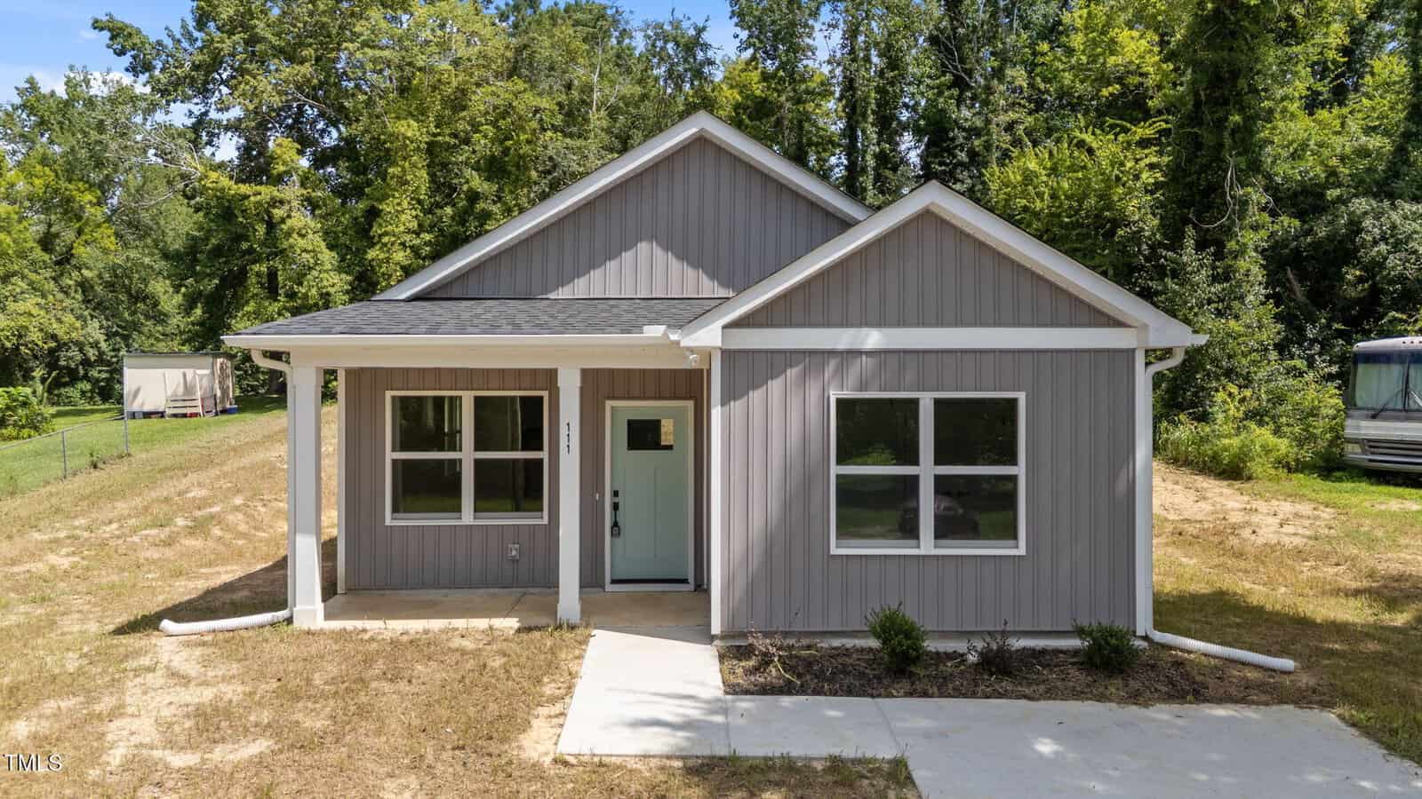 A small, single-story house with gray siding, white trim, and a covered front porch. There are two front windows and a light blue front door. The house is surrounded by grass and trees.