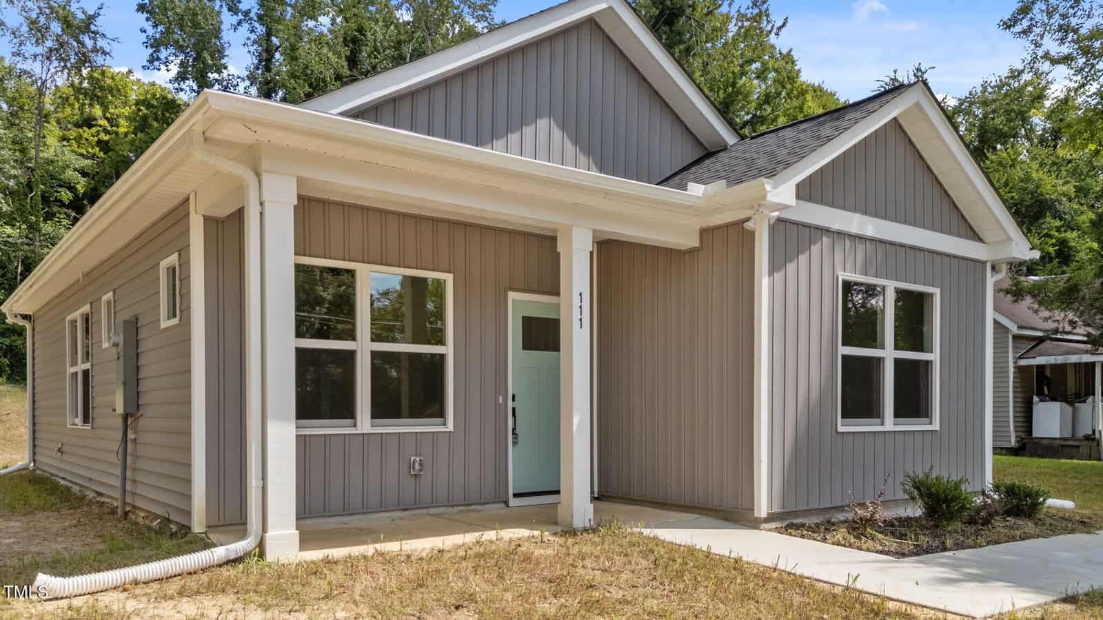 A single-story gray house with white trim, a covered front porch supported by two columns, a light blue front door, and several large windows. The house sits on a concrete walkway with sparse grass and trees in the background.
